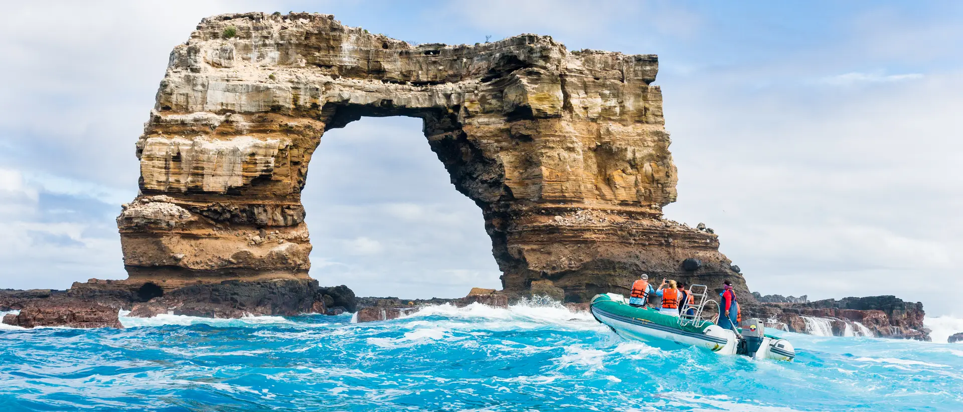 A boat with passengers approaches a large rock formation arching over turquoise waters under a cloudy sky.