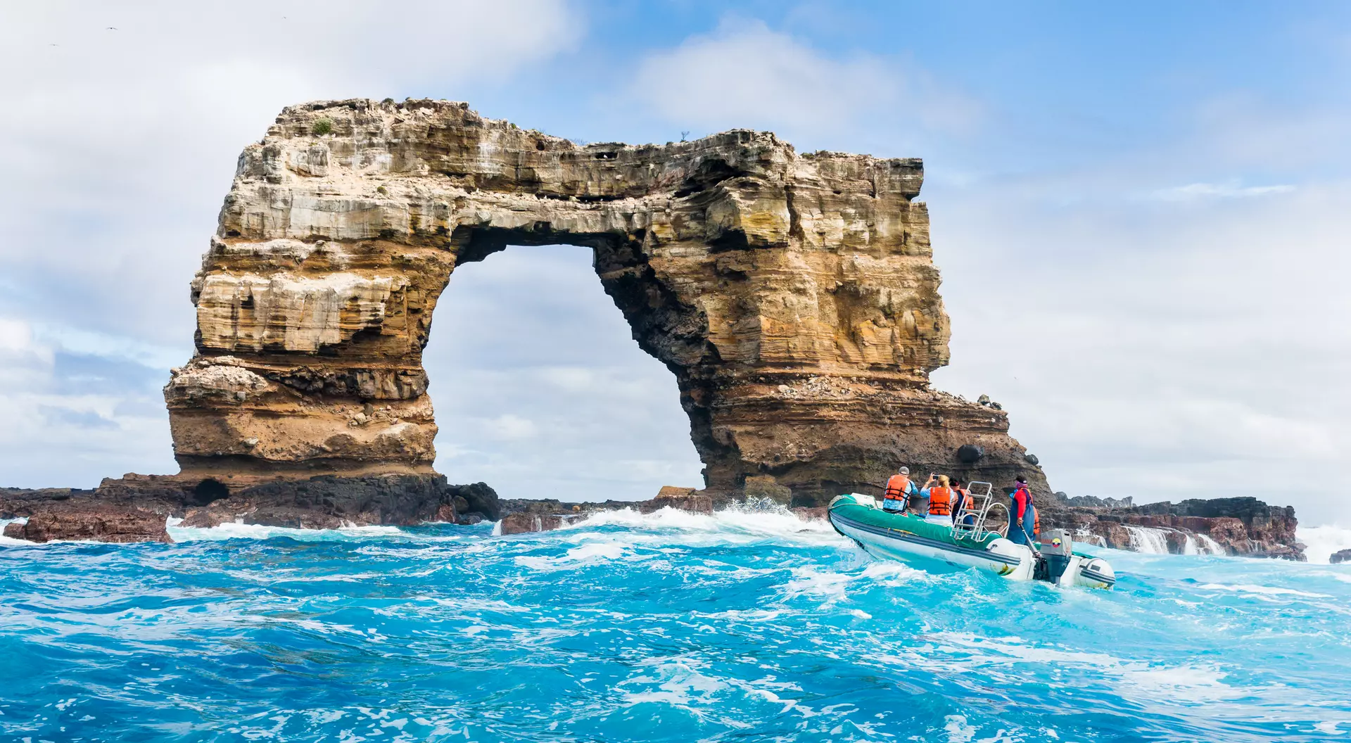 A boat with passengers approaches a large rock formation arching over turquoise waters under a cloudy sky.