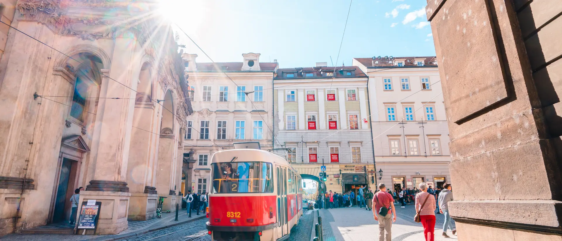 Red tram on a cobblestone street with sun shining, surrounded by historic buildings and people walking.