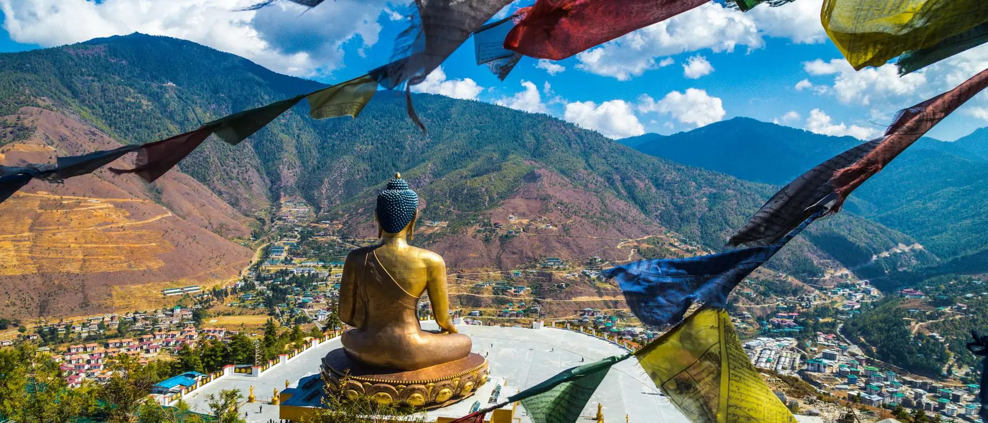 A large Buddha statue overlooks a valley, with colorful prayer flags fluttering in the foreground against a mountainous backdrop.