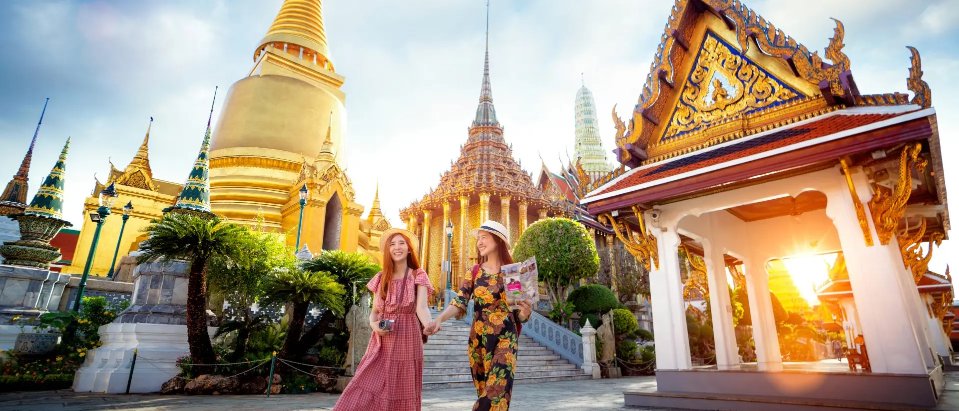 Two smiling women walk hand in hand, surrounded by ornate temples and golden spires, under a bright sky at sunset.