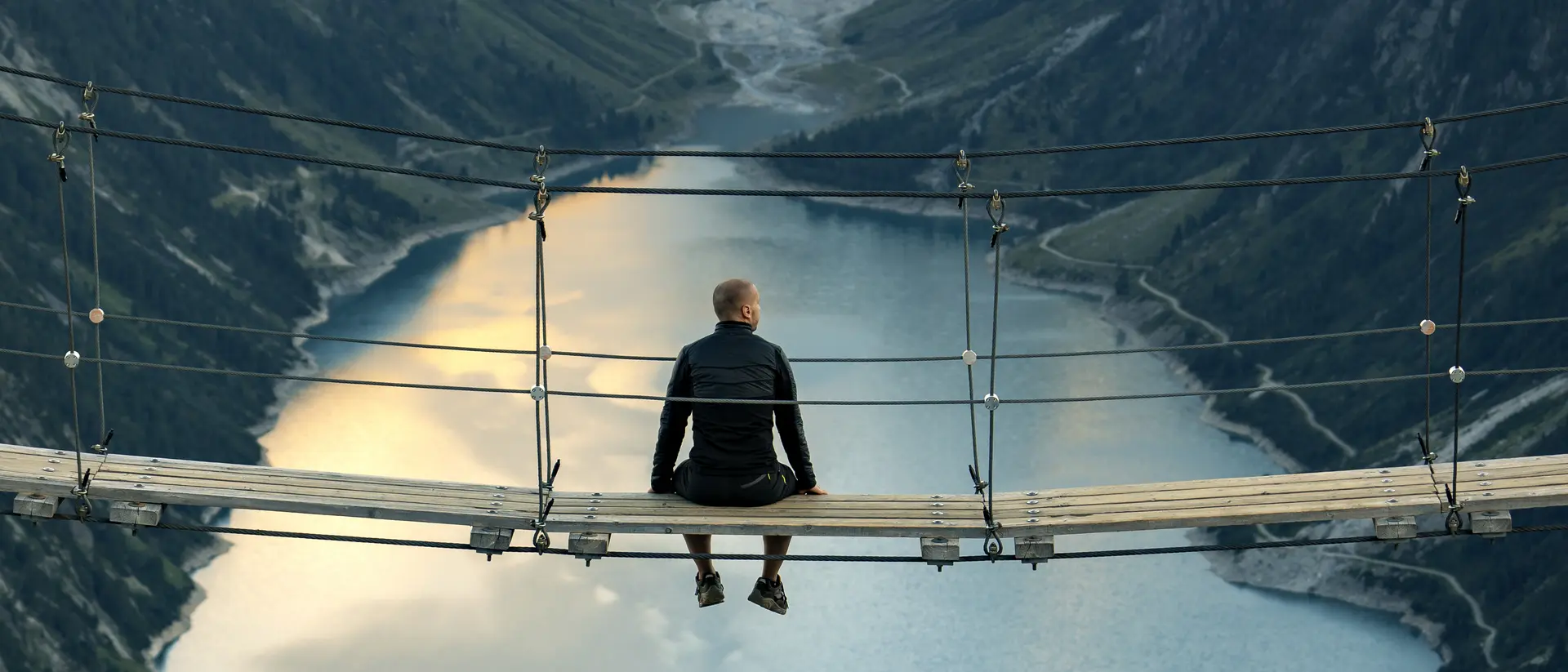 A person sitting on a wooden suspension bridge overlooking a deep valley and calm water below, with mountains in the background.