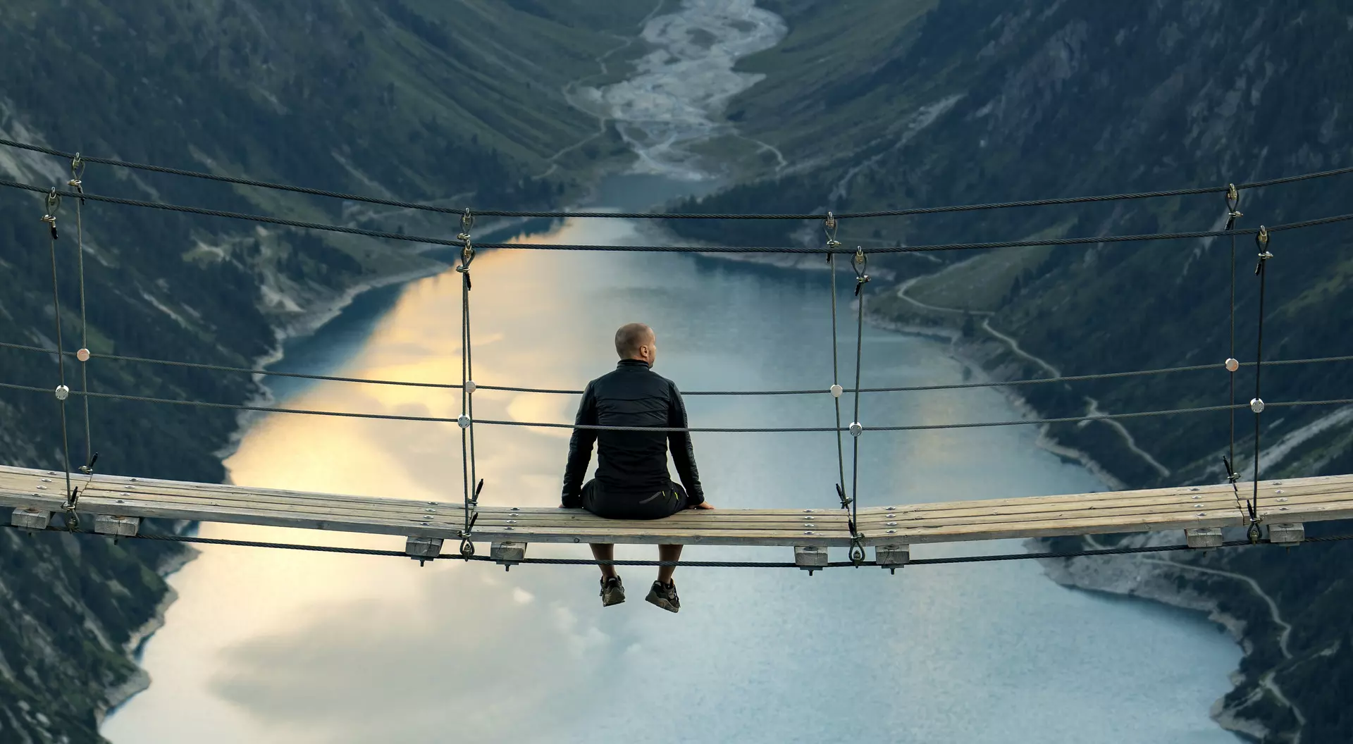 A person sitting on a wooden suspension bridge overlooking a deep valley and calm water below, with mountains in the background.