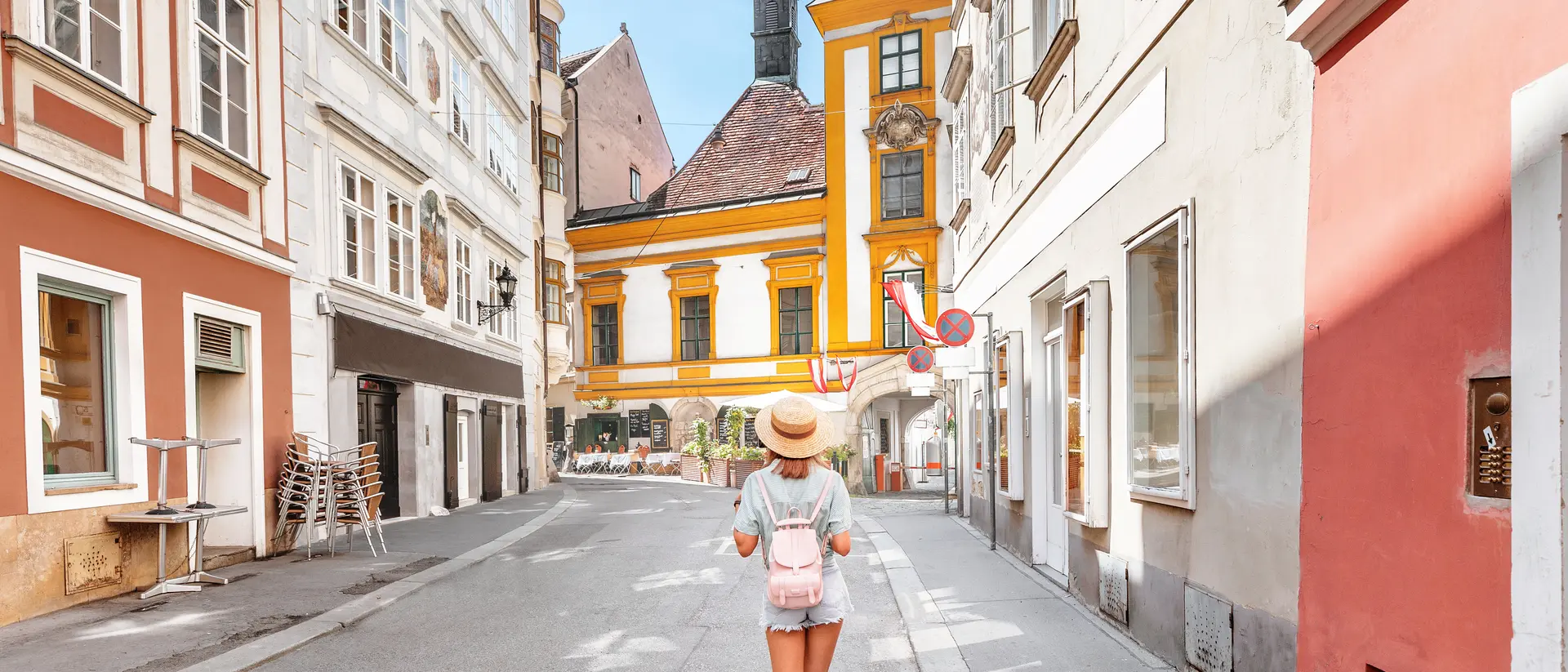 A person with a hat walks down a quaint street lined with colorful buildings and a church in the background.