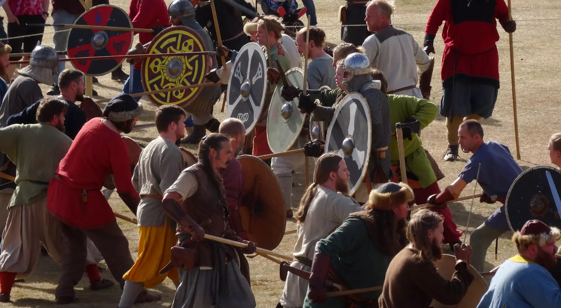A reenactment scene with people in medieval costumes wielding shields and weapons in a grassy area.
