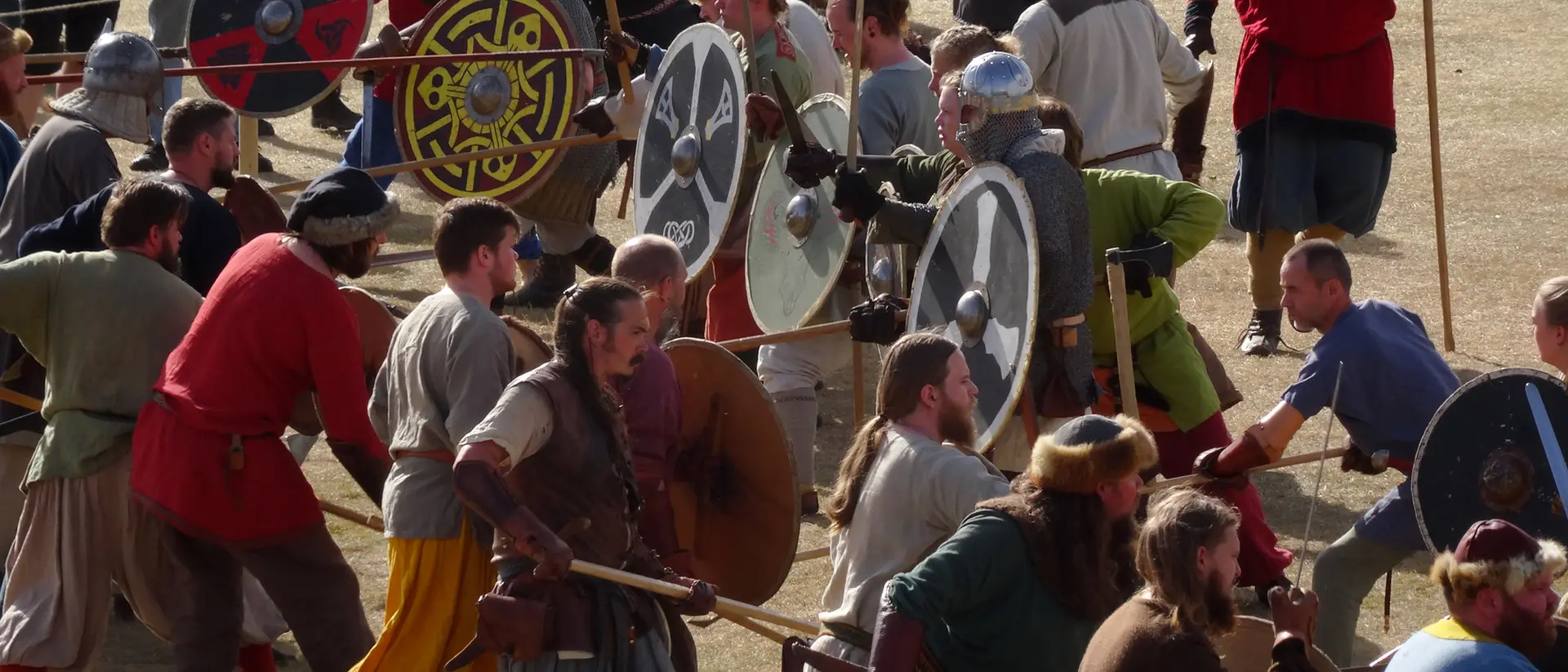 A reenactment scene with people in medieval costumes wielding shields and weapons in a grassy area.