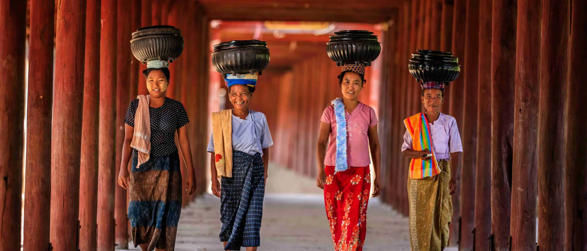Four girls walk confidently under a wooden archway, each balancing a container on their heads.