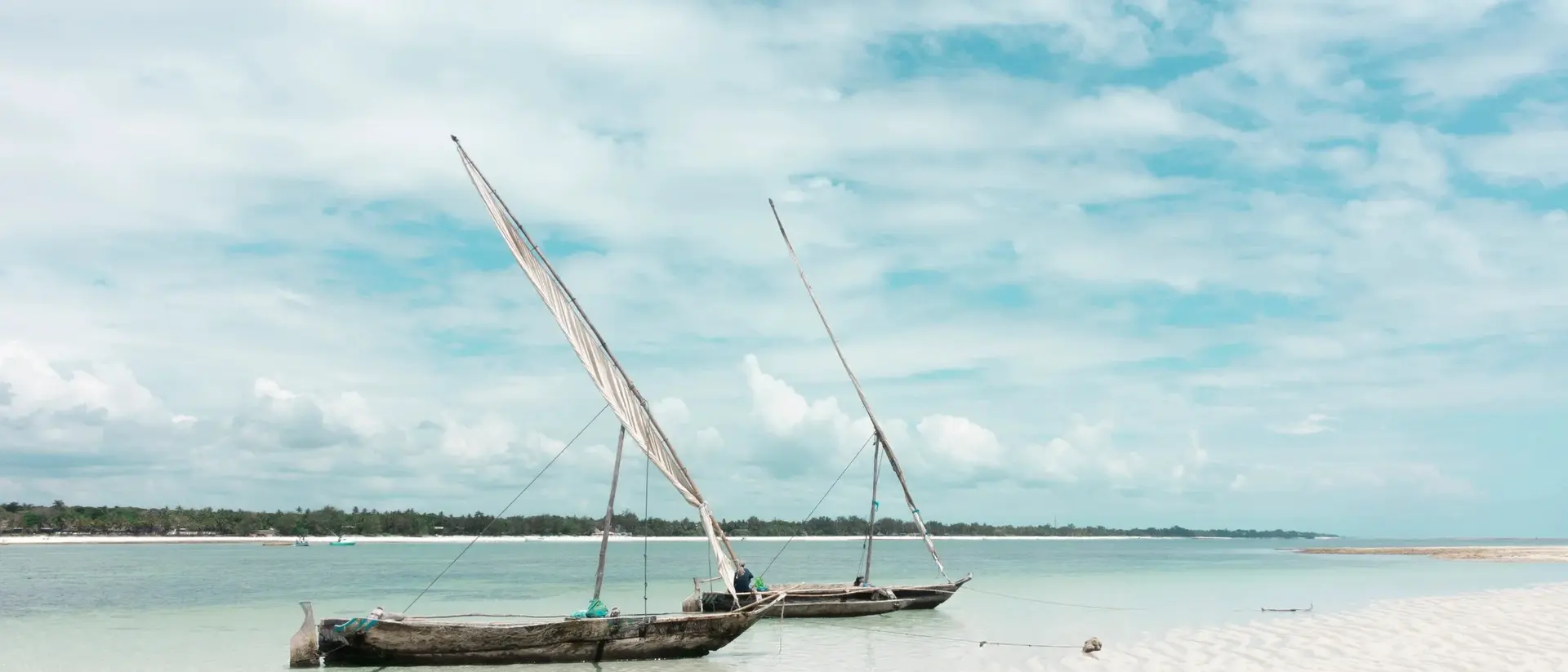 Two traditional sailboats on a calm beach, with soft sand and a cloudy sky in the background.