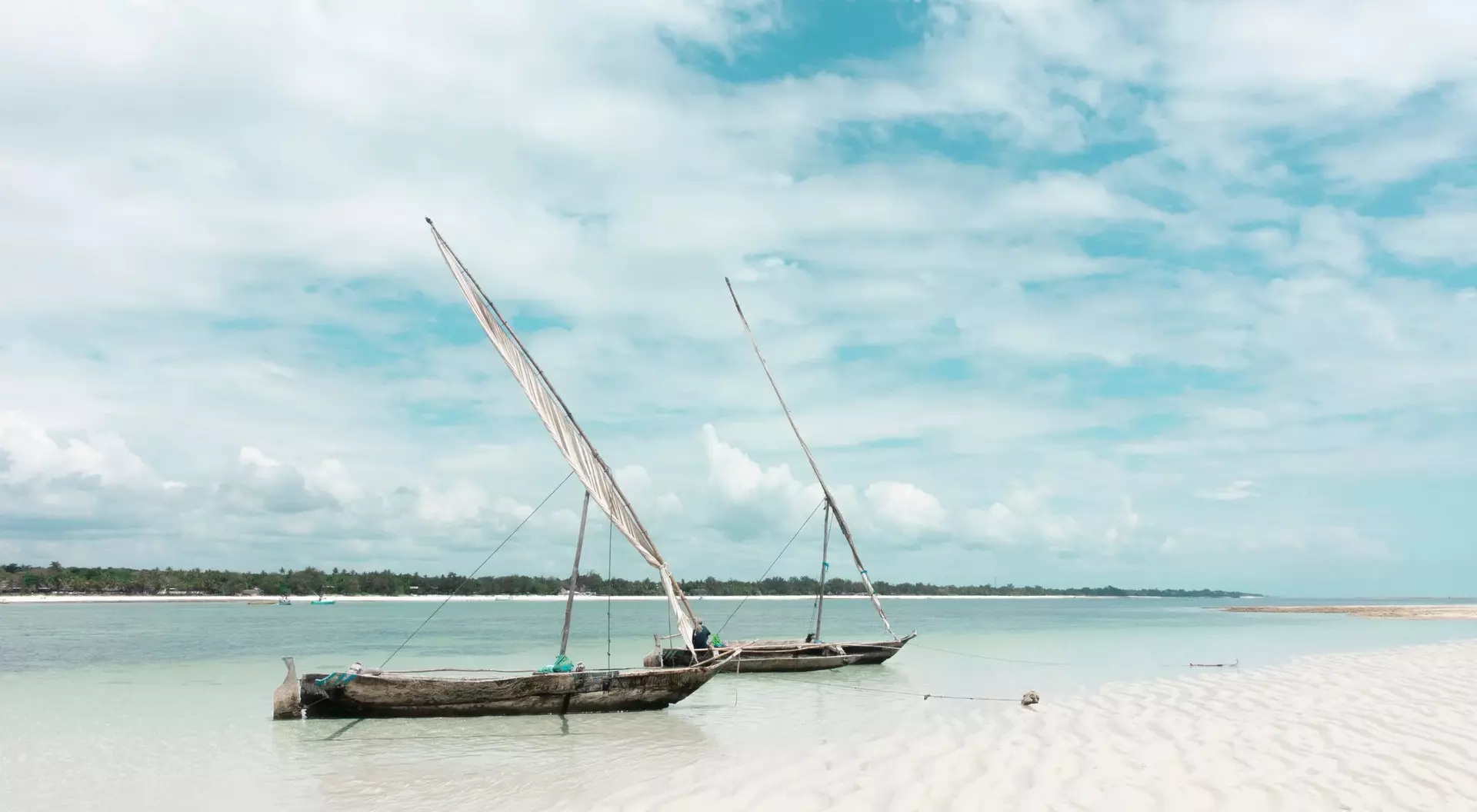 Two traditional sailboats on a calm beach, with soft sand and a cloudy sky in the background.