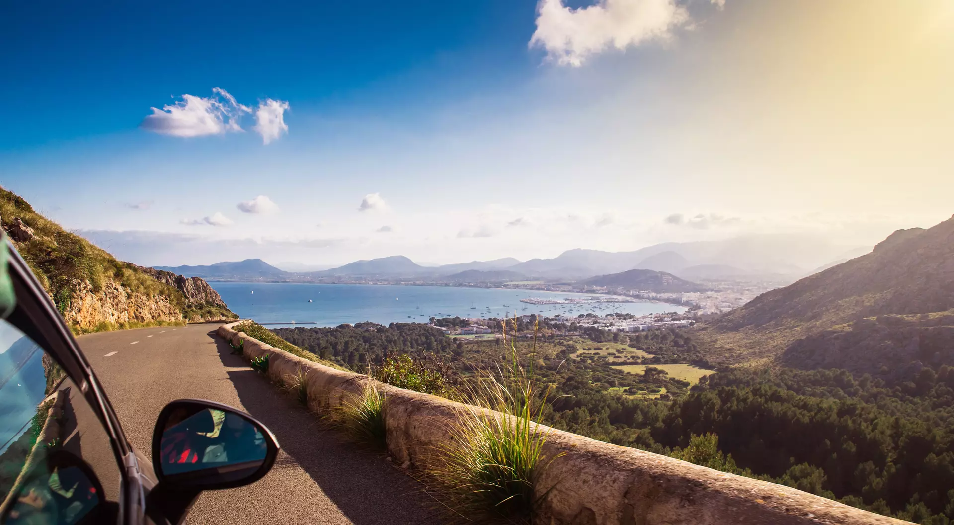 A scenic view from a car overlooking a coastline, mountains, and blue sky with clouds. Sunlight shines brightly.