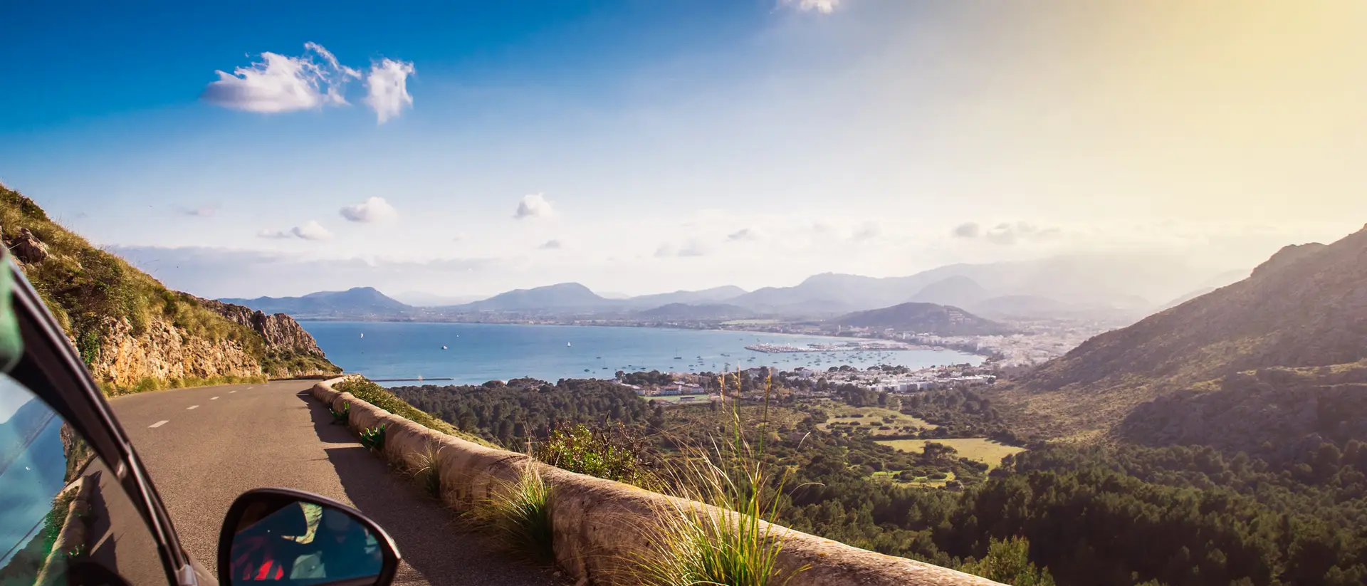 A scenic view from a car overlooking a coastline, mountains, and blue sky with clouds. Sunlight shines brightly.