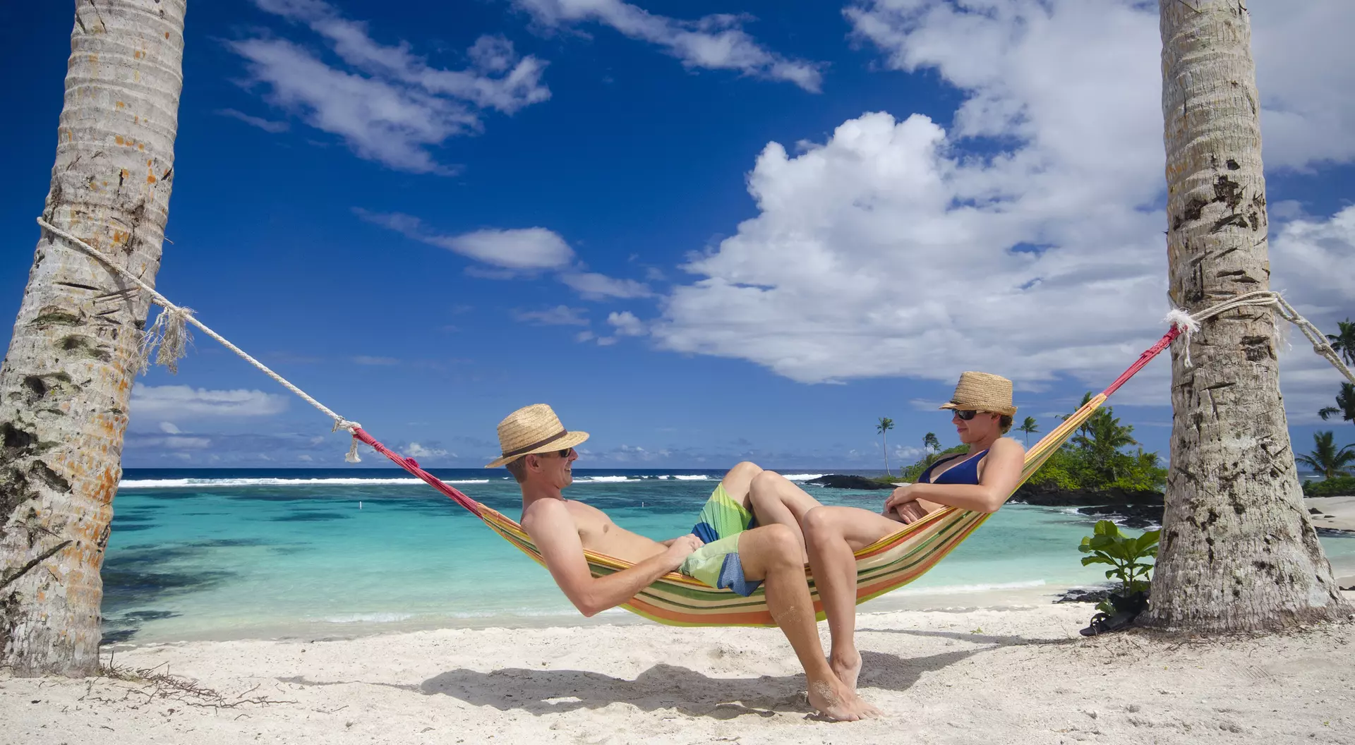 Two people relax in a hammock between palm trees on a sandy beach, with a clear blue ocean and sky in the background.