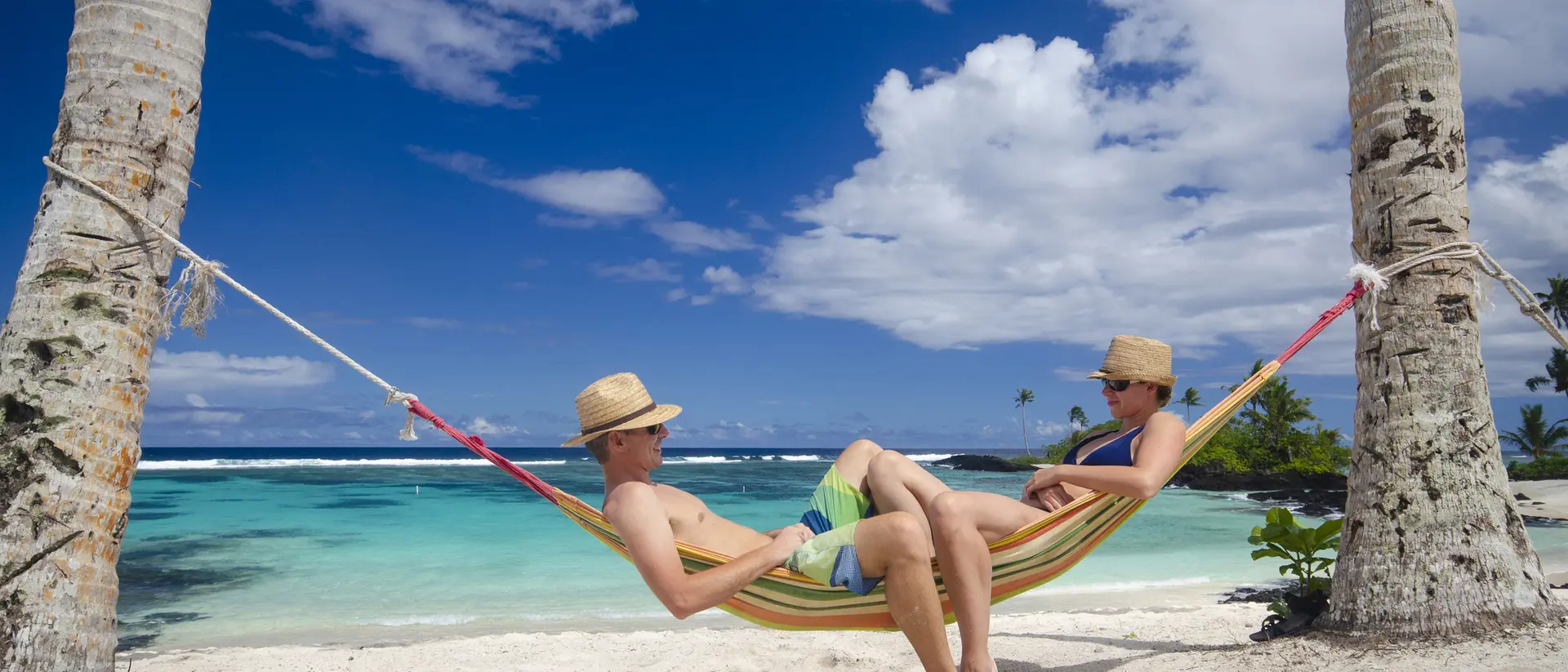 Two people relax in a hammock between palm trees on a sandy beach, with a clear blue ocean and sky in the background.