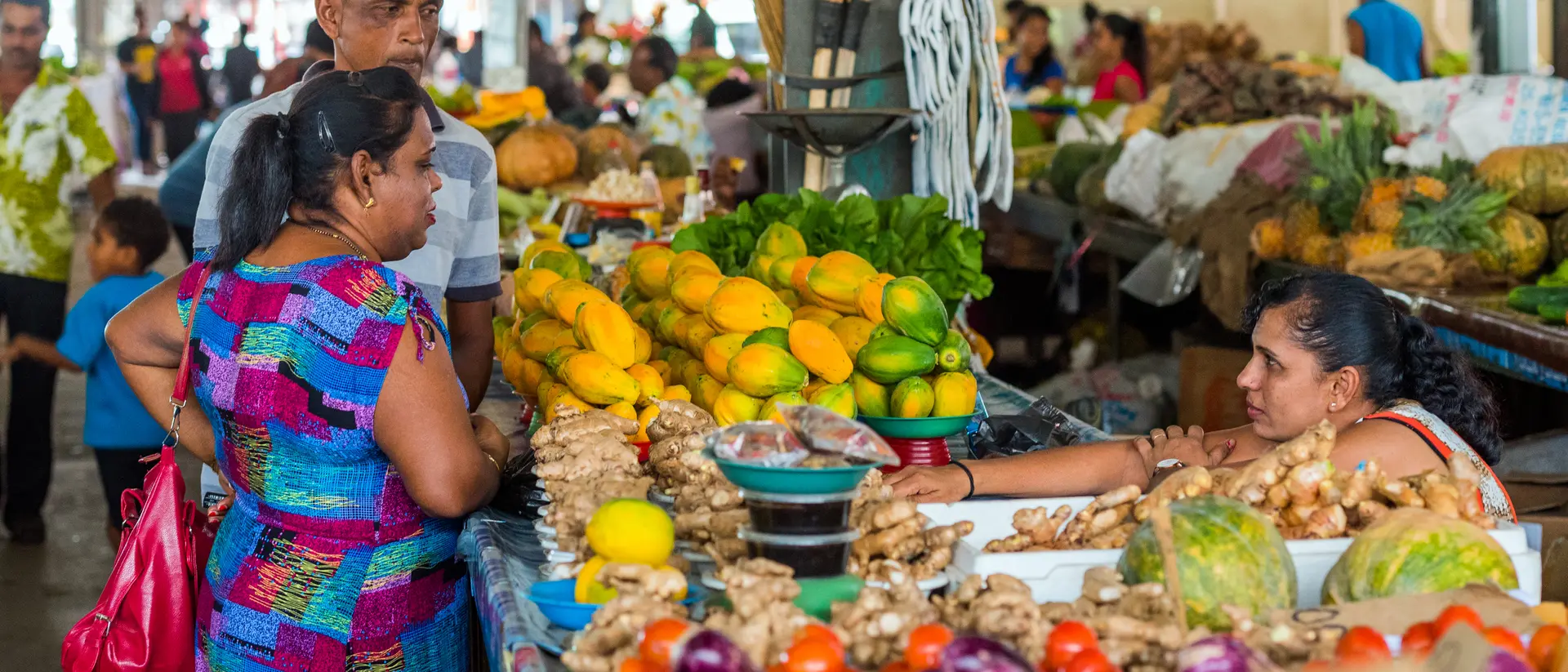 Two women and a man shop at a vibrant market, surrounded by fresh fruits and vegetables in a lively atmosphere.