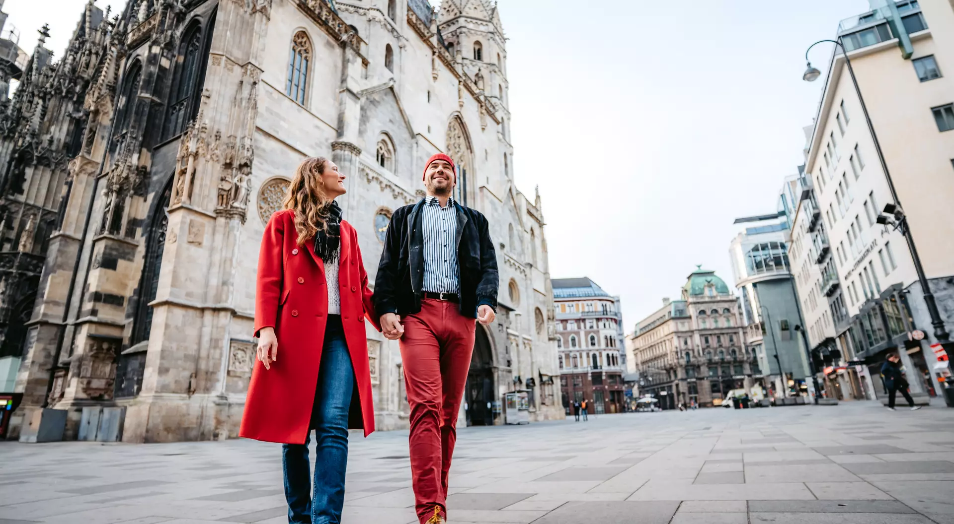 A couple walks hand in hand through a city square, with historical architecture in the background.