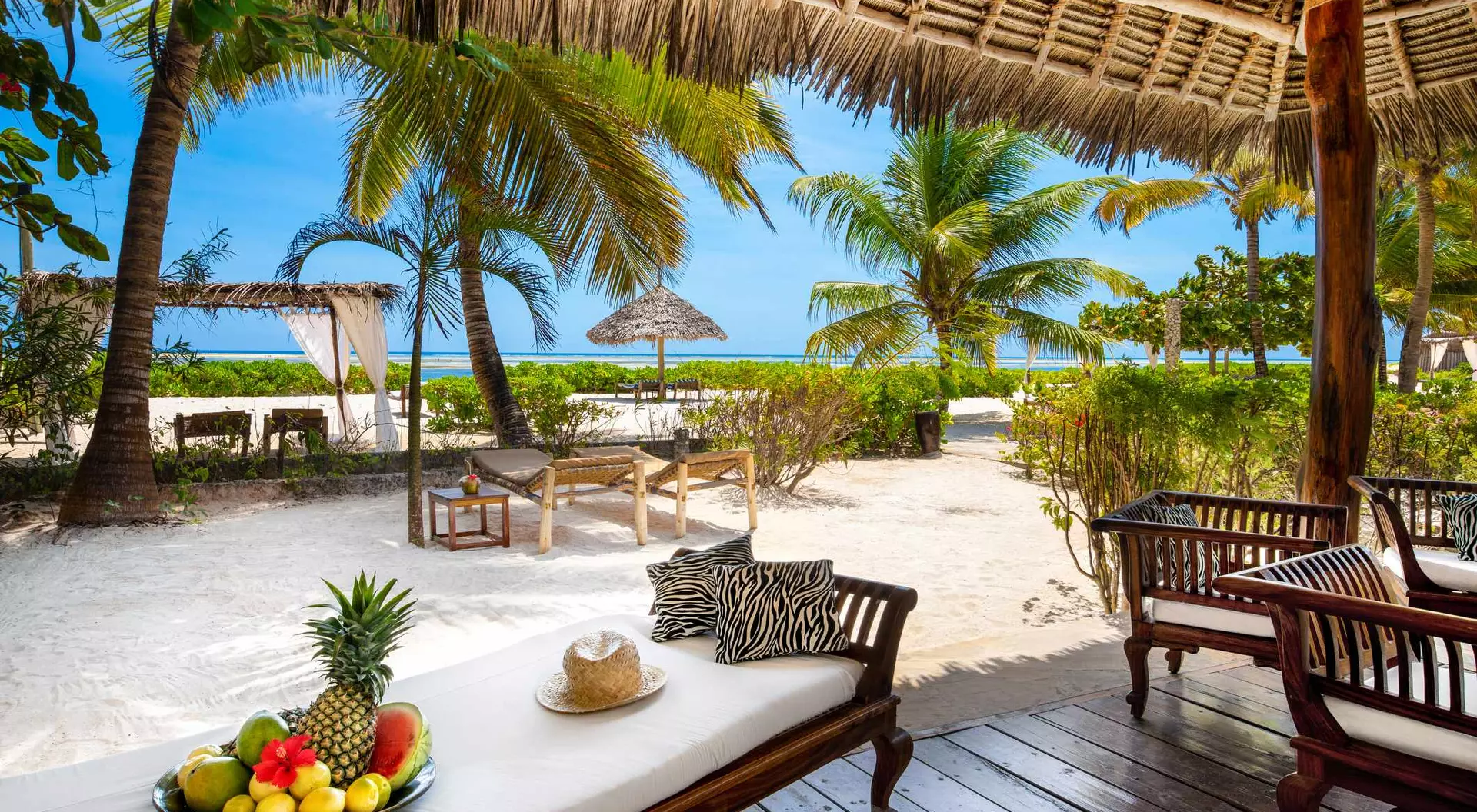 Tropical beach scene with palm trees, a fruit platter, lounge chairs, and a thatched roof against a blue sky.