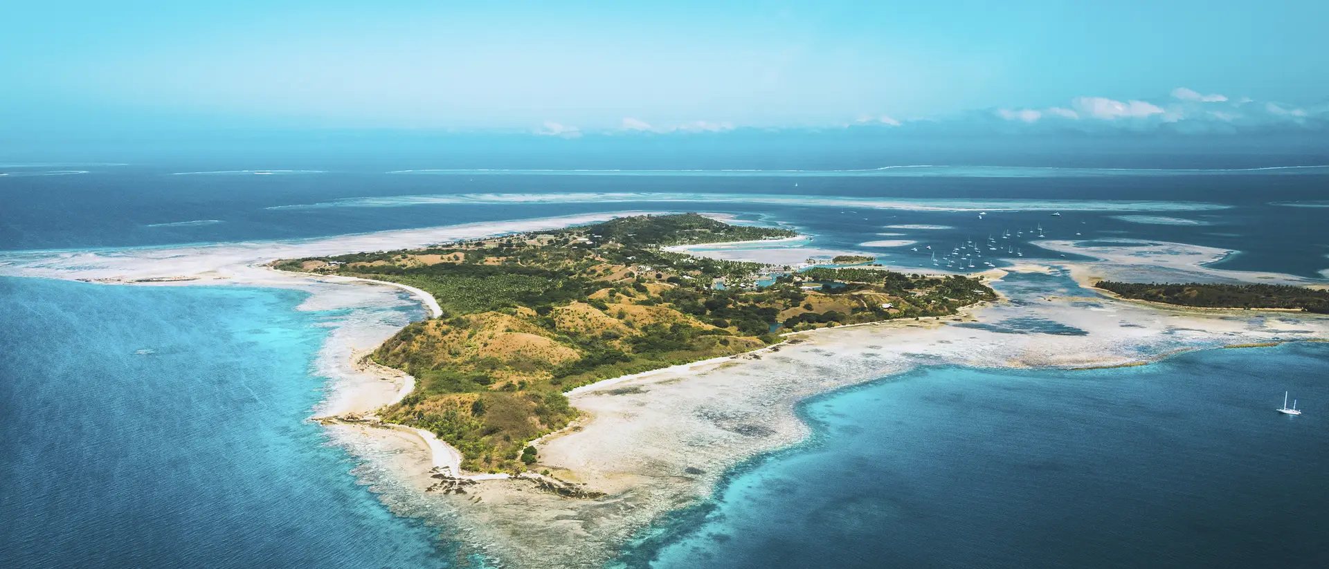 Aerial view of a small, lush island surrounded by turquoise water and coral reefs under a clear blue sky.