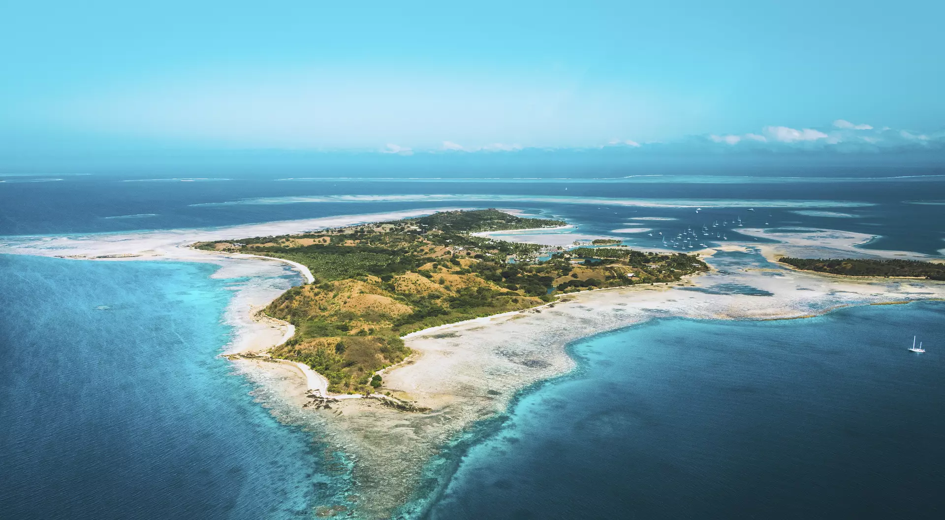 Aerial view of a small, lush island surrounded by turquoise water and coral reefs under a clear blue sky.