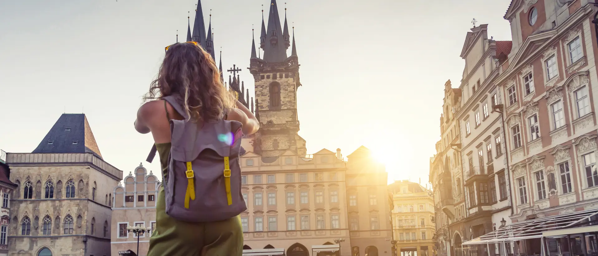 A person with a backpack stands in an empty square, gazing at historic buildings under a golden sunset.