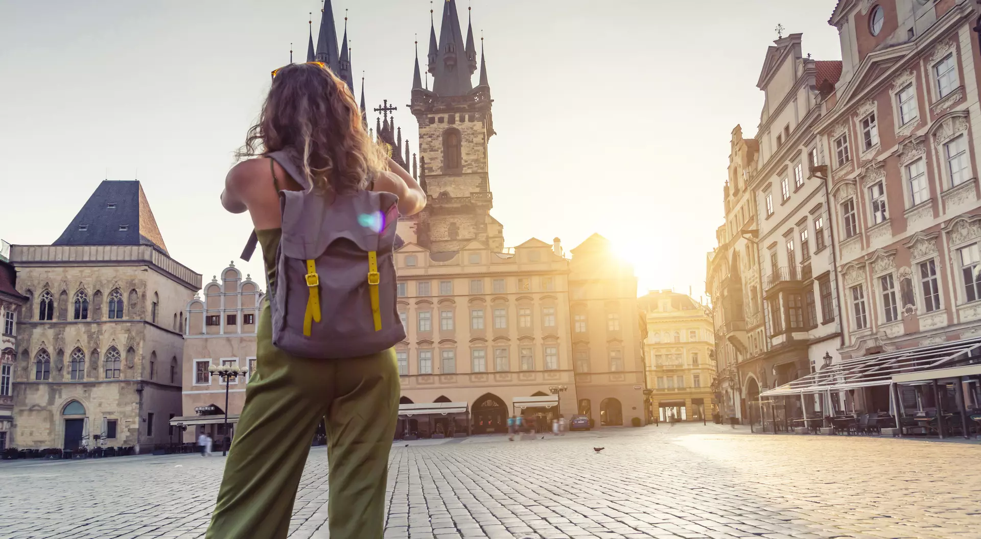 A person with a backpack stands in an empty square, gazing at historic buildings under a golden sunset.