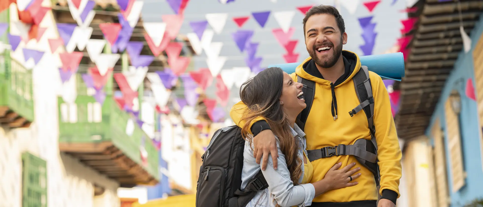A joyful couple with backpacks laugh together in a colorful street decorated with festive flags.