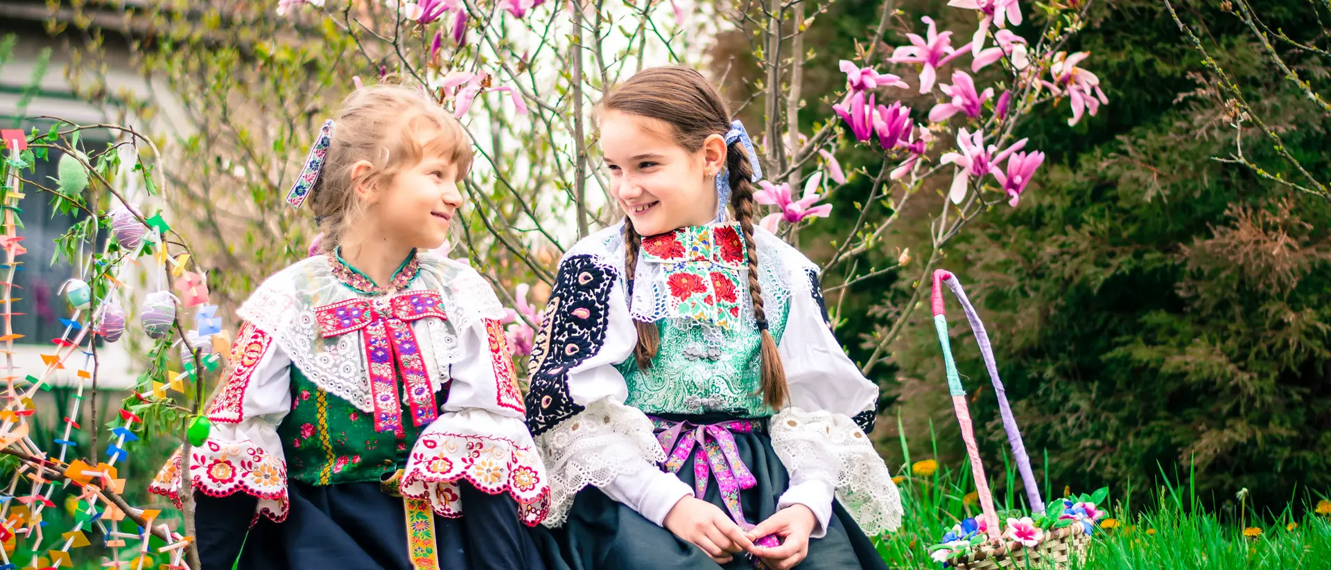 Two girls in traditional costumes sit on the grass, smiling among blooming flowers and plants.