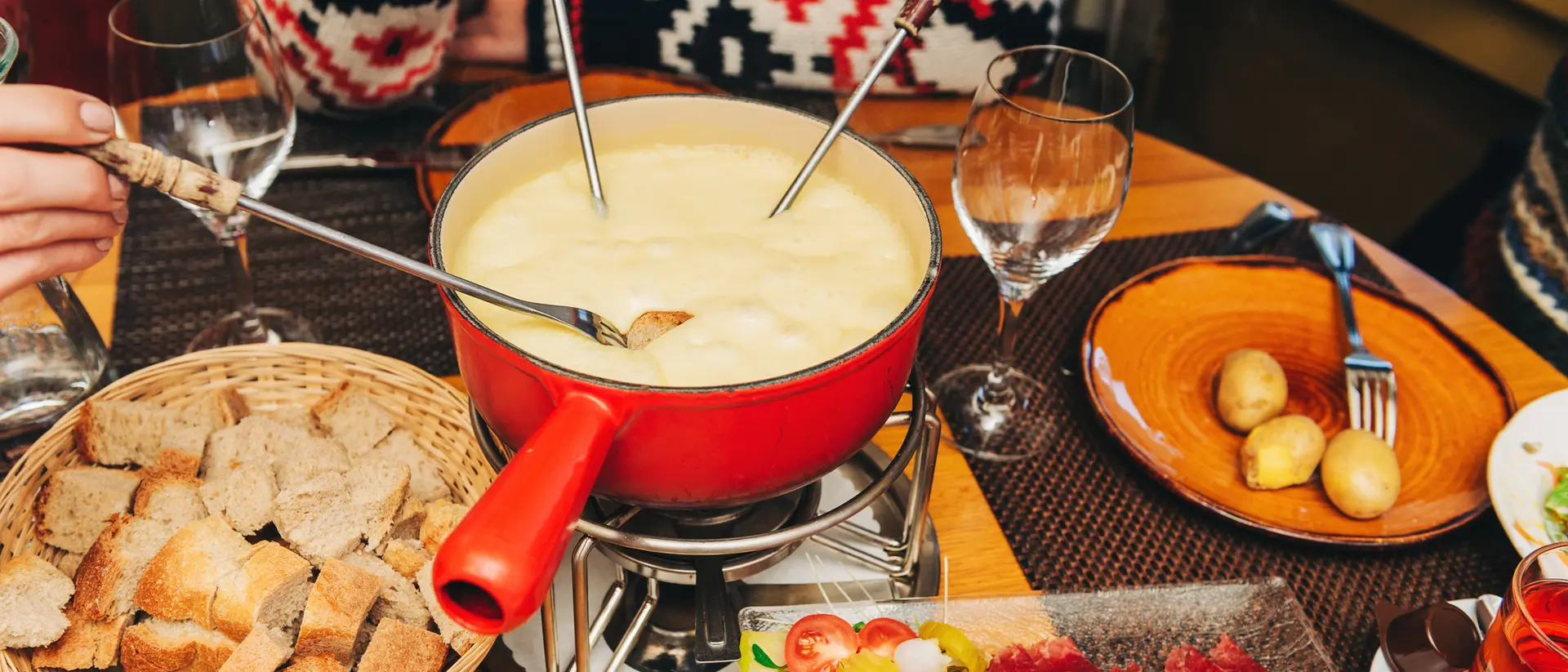 A cheese fondue pot surrounded by bread, vegetables, and drinks, with hands dipping into the fondue.