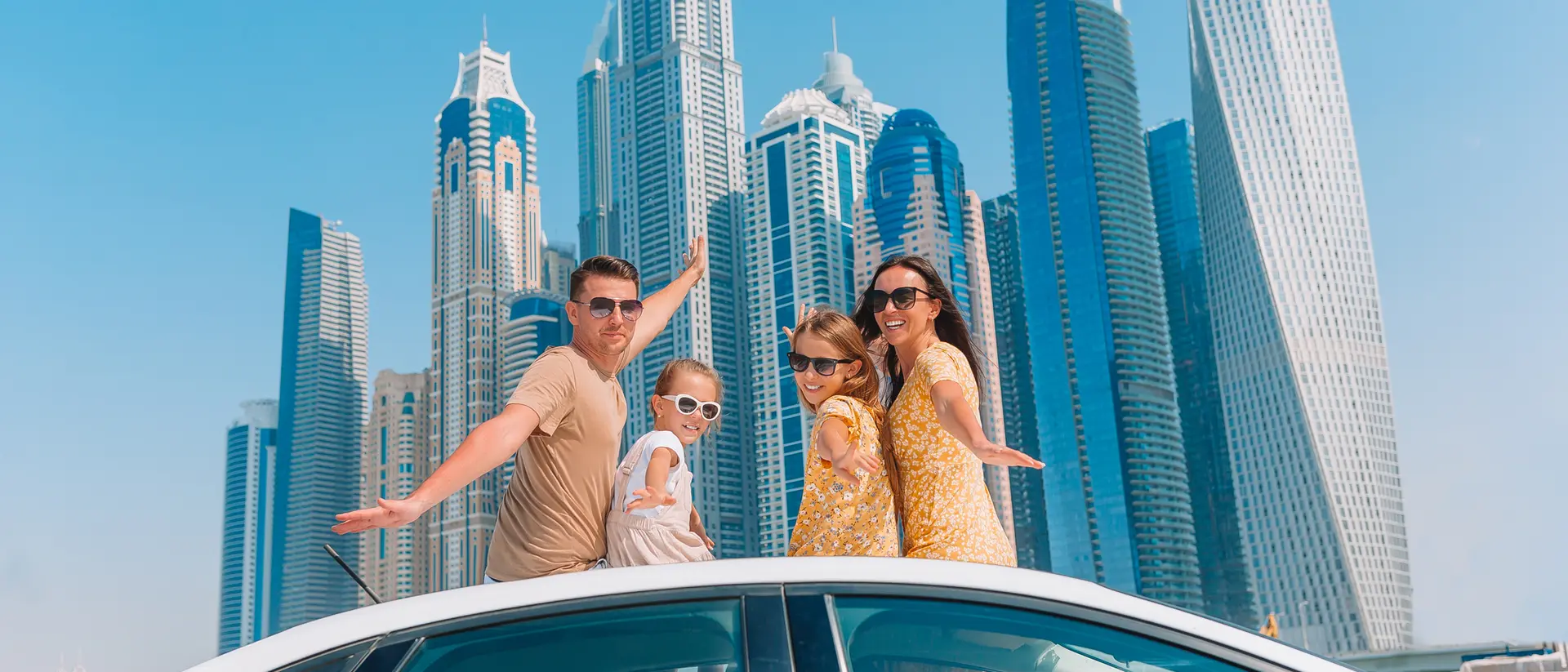 A family poses on a car rooftop with skyscrapers in the background on a sunny day.