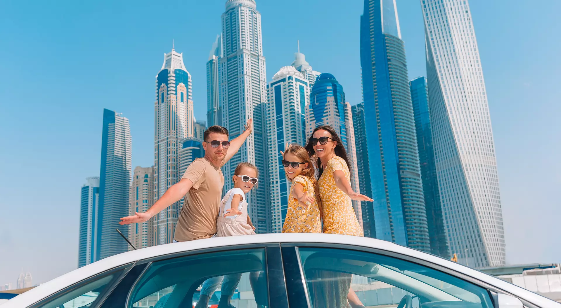 A family poses on a car rooftop with skyscrapers in the background on a sunny day.