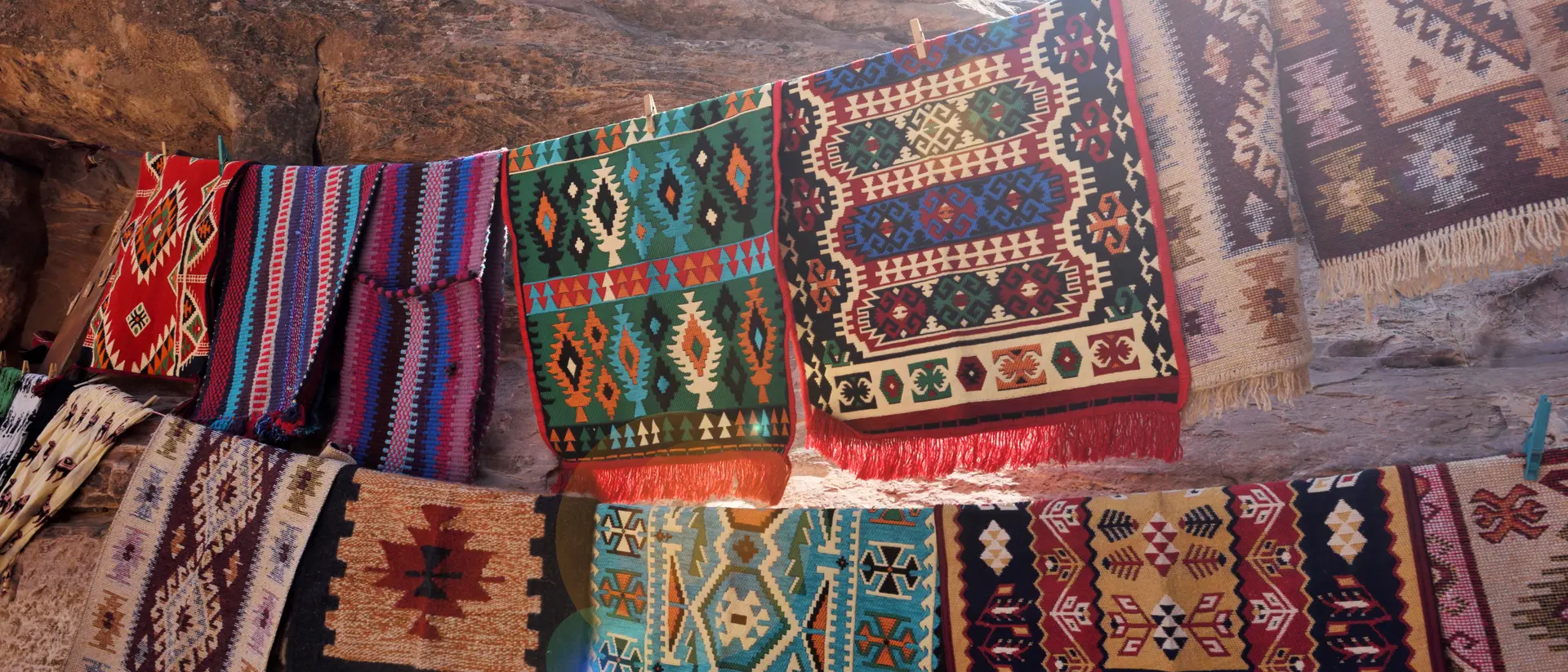 Colorful woven rugs displayed on a rocky surface, showcasing intricate patterns and textures in sunlight.