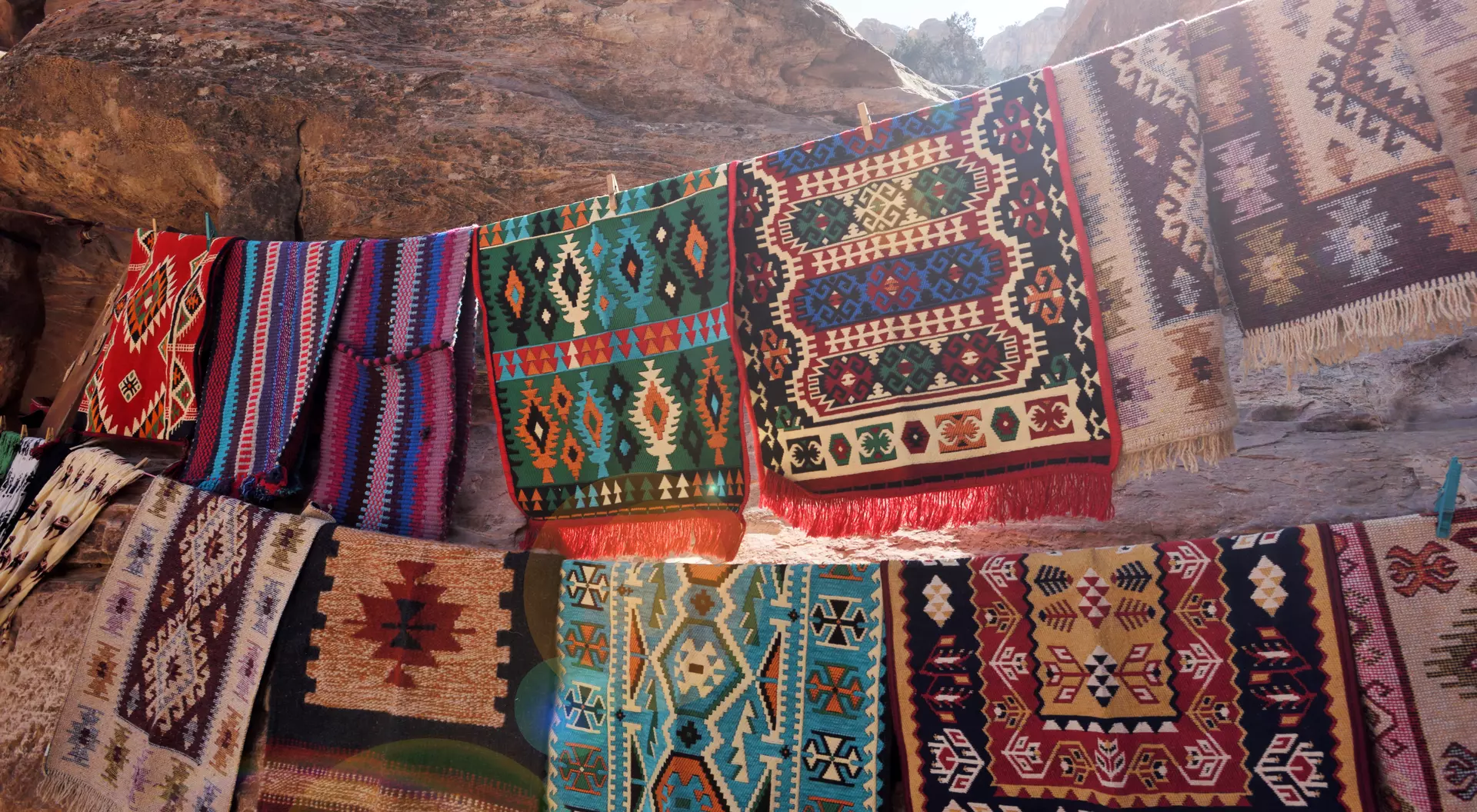 Colorful woven rugs displayed on a rocky surface, showcasing intricate patterns and textures in sunlight.