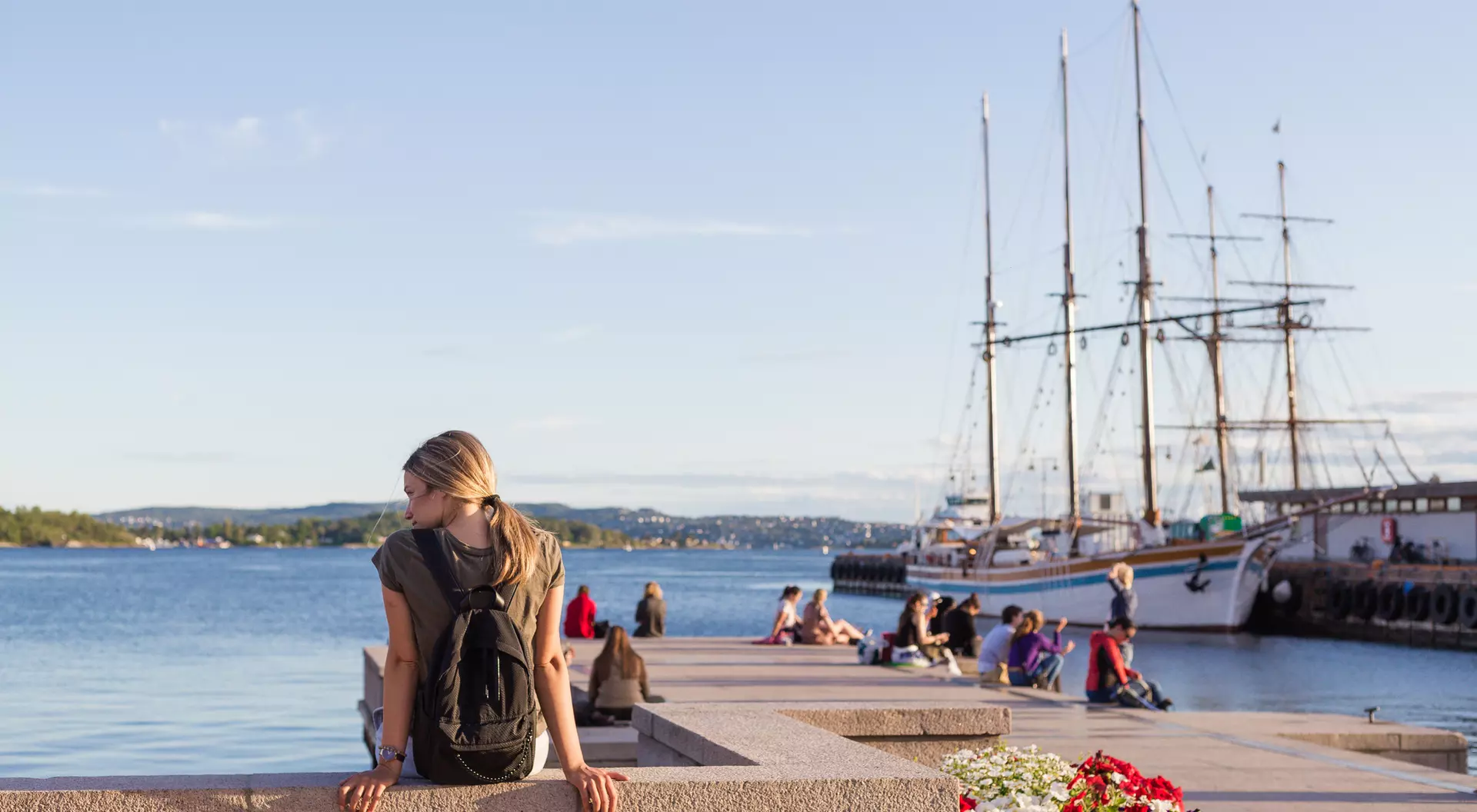 A woman with a backpack sits on a waterfront wall, looking at the water and nearby ships, with people and flowers around.