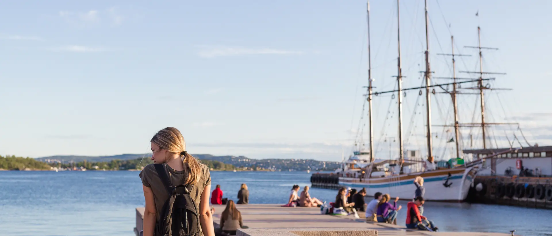 A woman with a backpack sits on a waterfront wall, looking at the water and nearby ships, with people and flowers around.