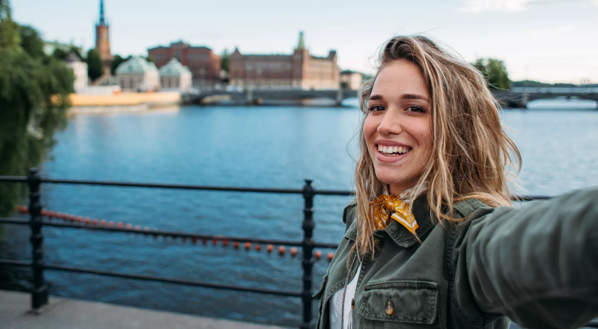 A smiling person takes a selfie by a waterfront with buildings and a spire in the background under a clear blue sky.