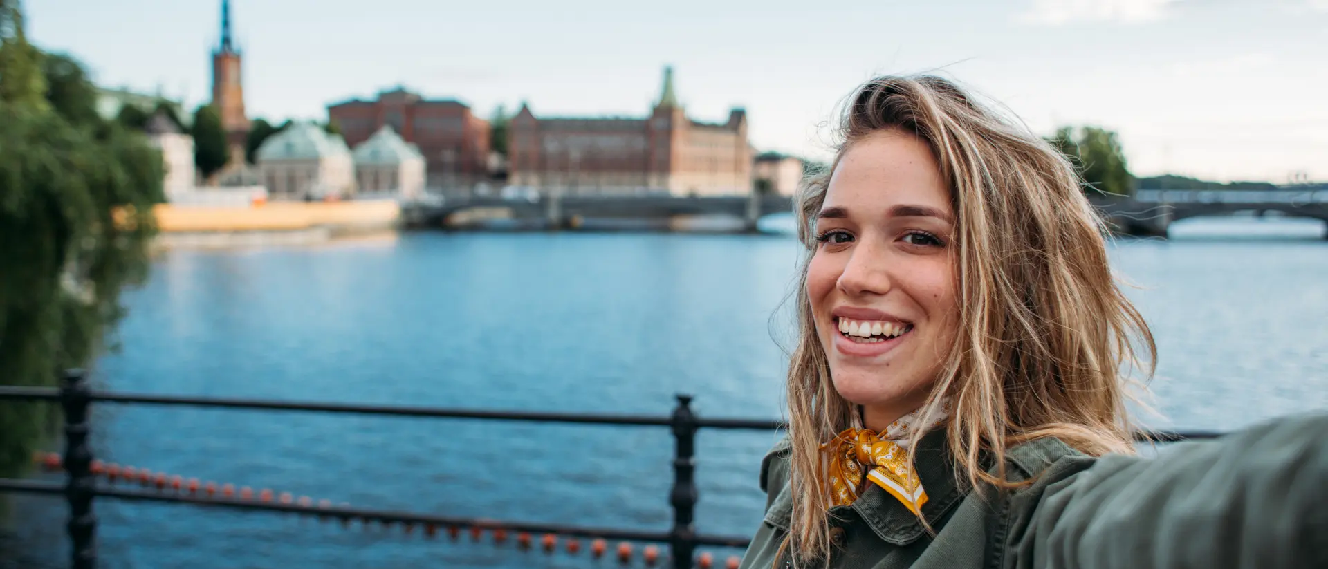 A smiling person takes a selfie by a waterfront with buildings and a spire in the background under a clear blue sky.