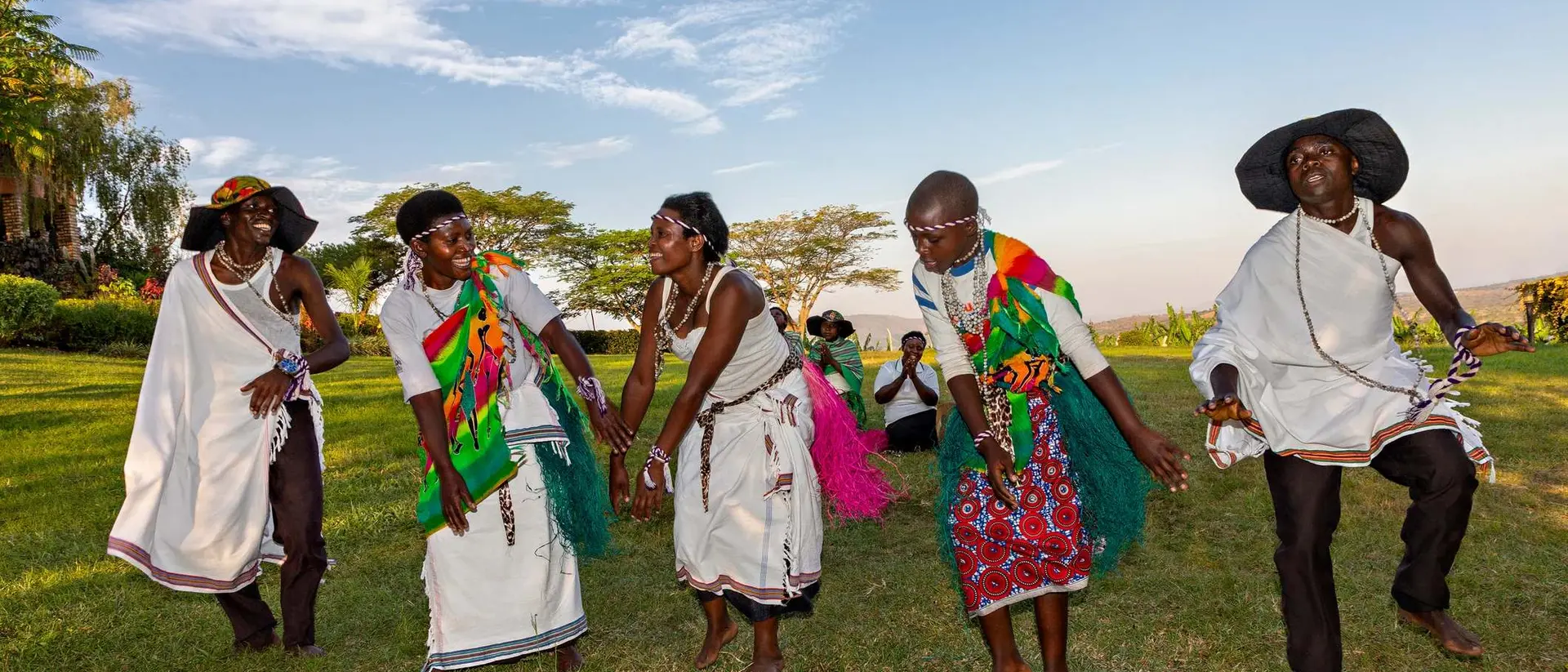 Group of dancers in colorful traditional attire performing outdoors, with a scenic landscape in the background.