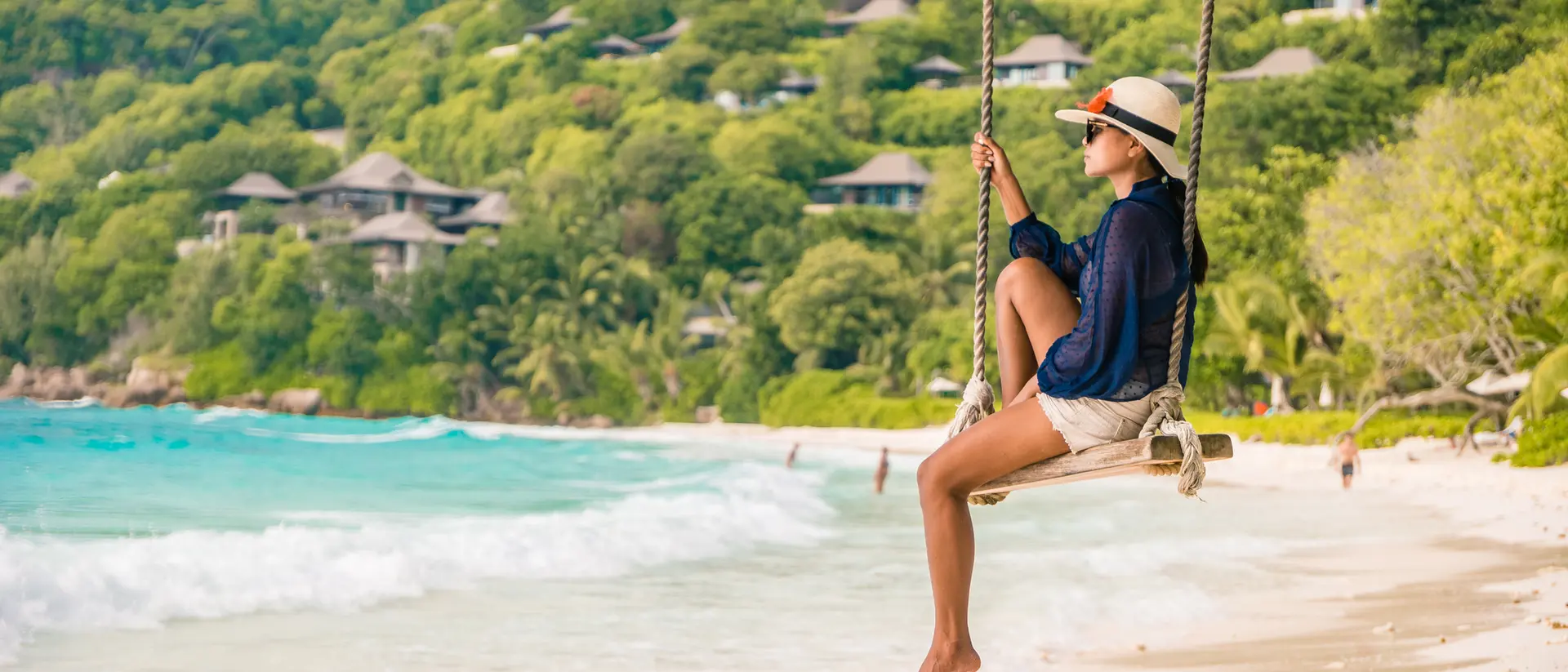 A woman sits on a swing by the beach, with turquoise waves and lush green hills in the background.