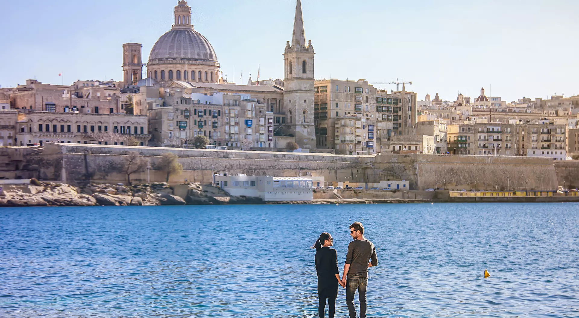 A couple stands hand in hand by the seaside, with a historic city skyline and dome in the background.