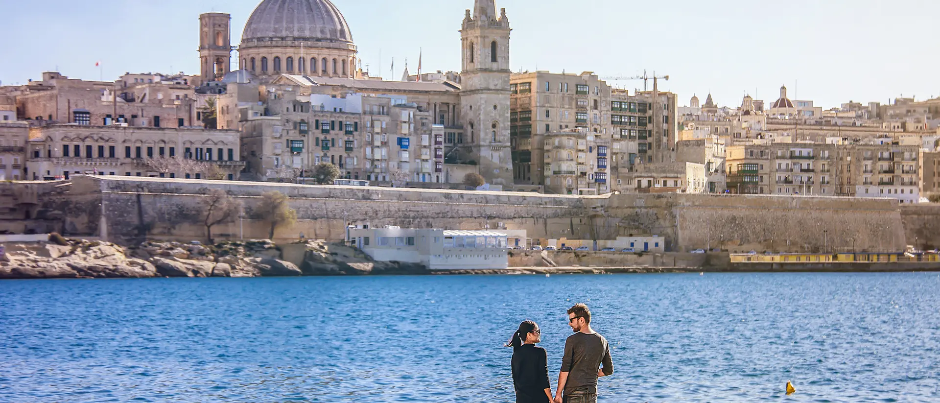 A couple stands hand in hand by the seaside, with a historic city skyline and dome in the background.