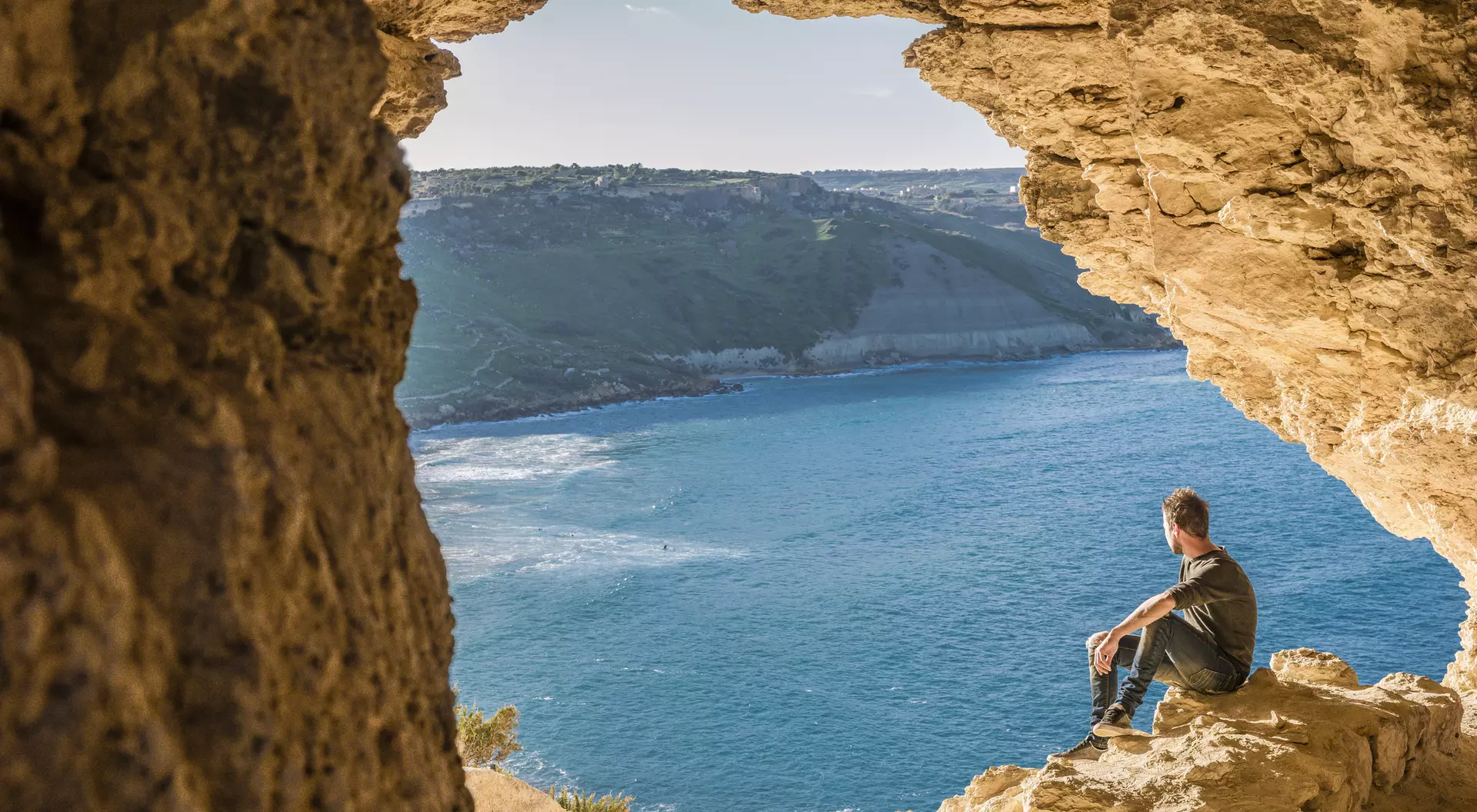 A person sits on rocky ledge, gazing at the ocean view from a cave entrance.