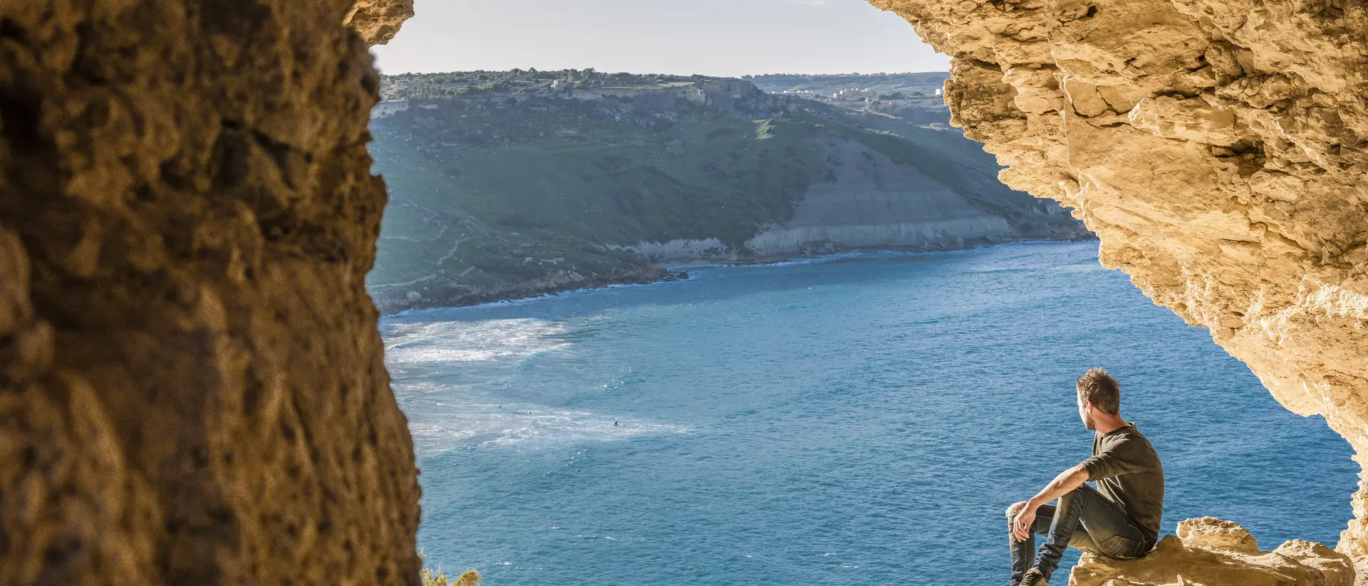 A person sits on rocky ledge, gazing at the ocean view from a cave entrance.