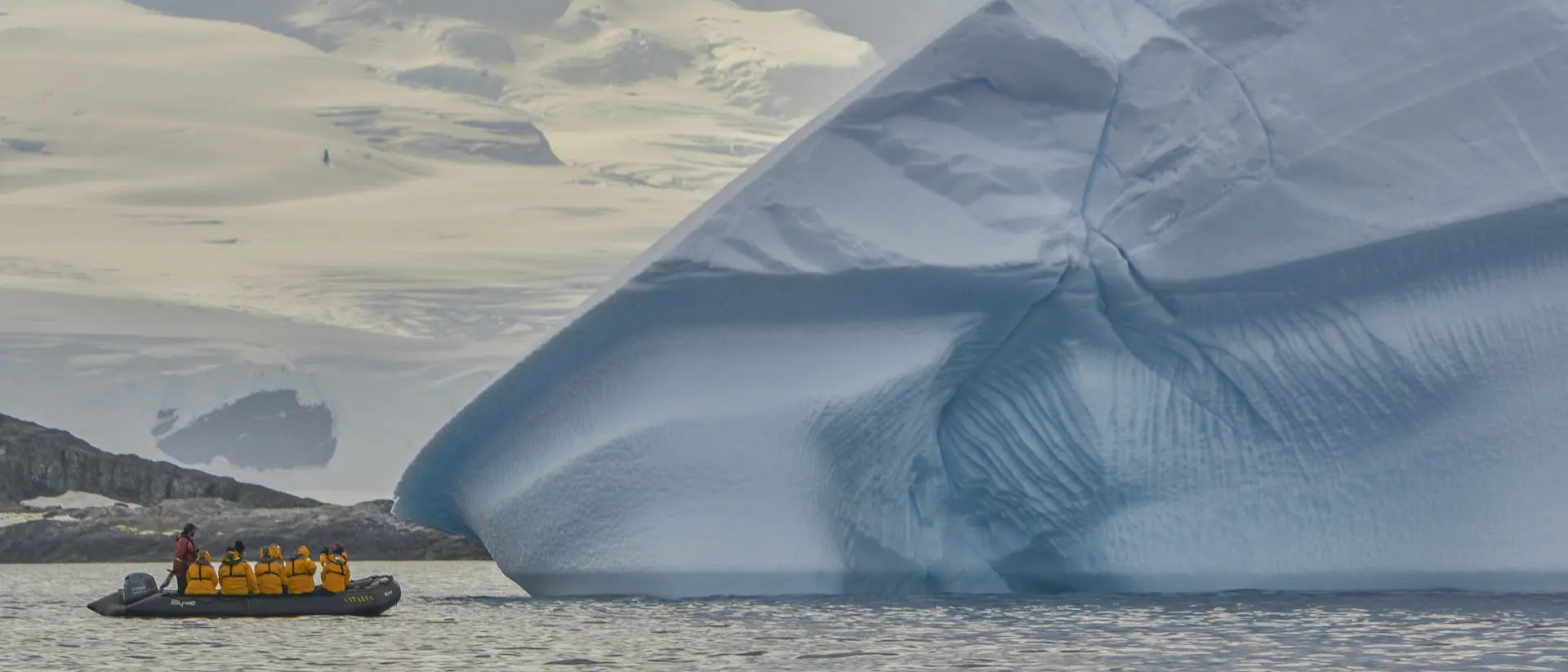 A small boat with passengers approaches a large blue iceberg in a calm sea, surrounded by icy landscapes and clouds.