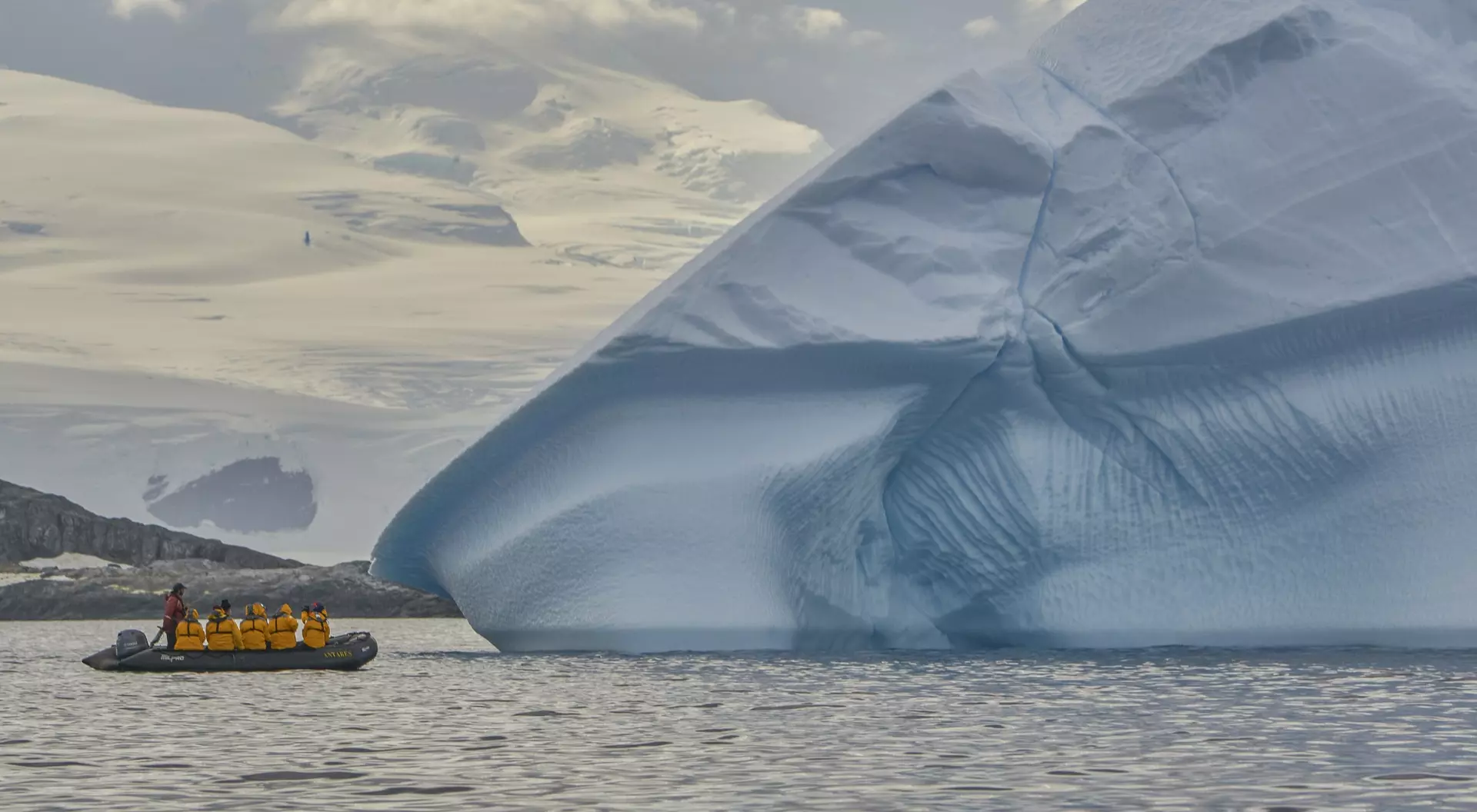 A small boat with passengers approaches a large blue iceberg in a calm sea, surrounded by icy landscapes and clouds.