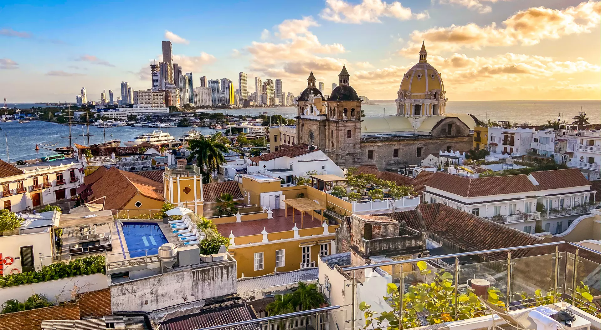A panoramic view of Cartagena, Colombia, featuring colorful rooftops, a marina, and modern buildings against a sunset sky.