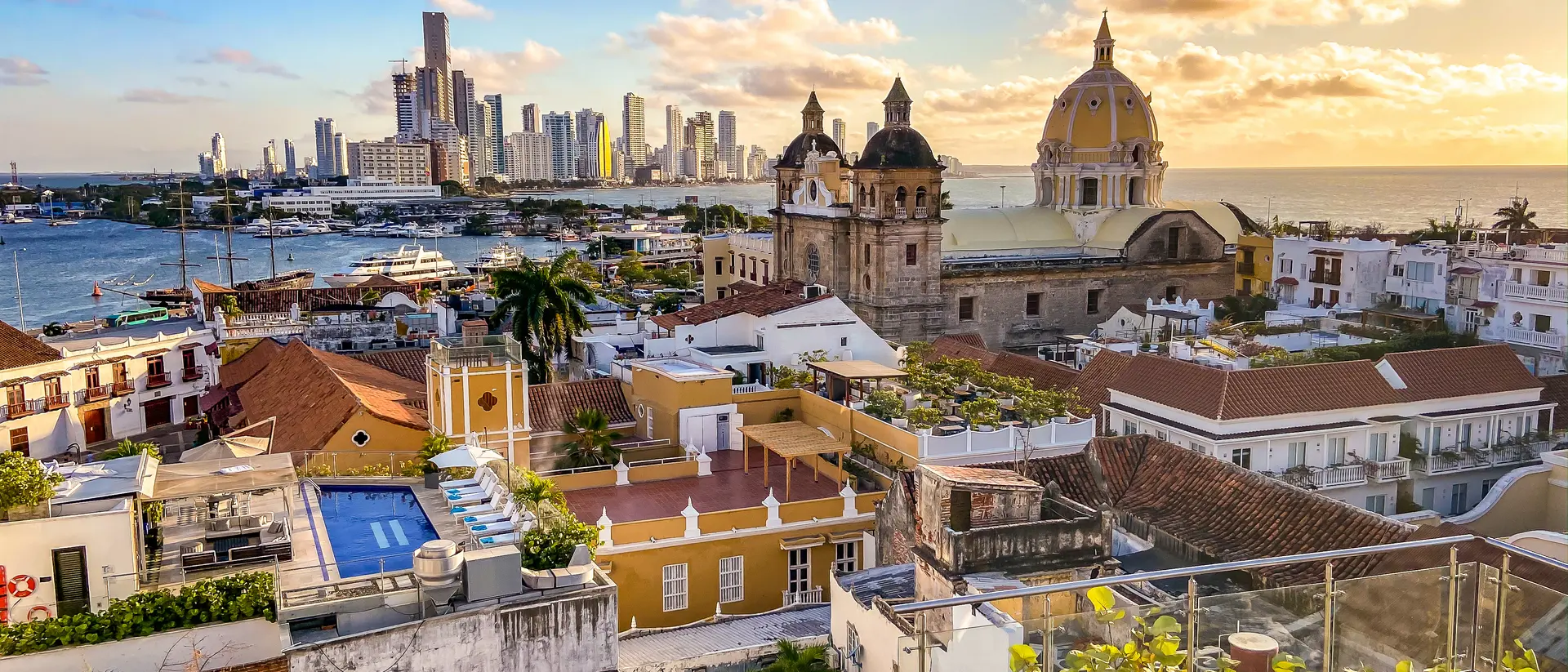 A panoramic view of Cartagena, Colombia, featuring colorful rooftops, a marina, and modern buildings against a sunset sky.