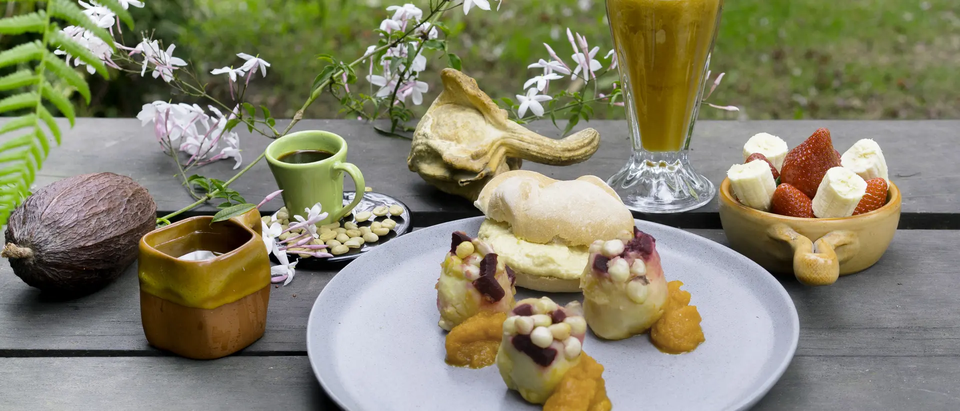 A plate of colorful desserts with a drink, surrounded by flowers and chocolate ingredients on a wooden table.