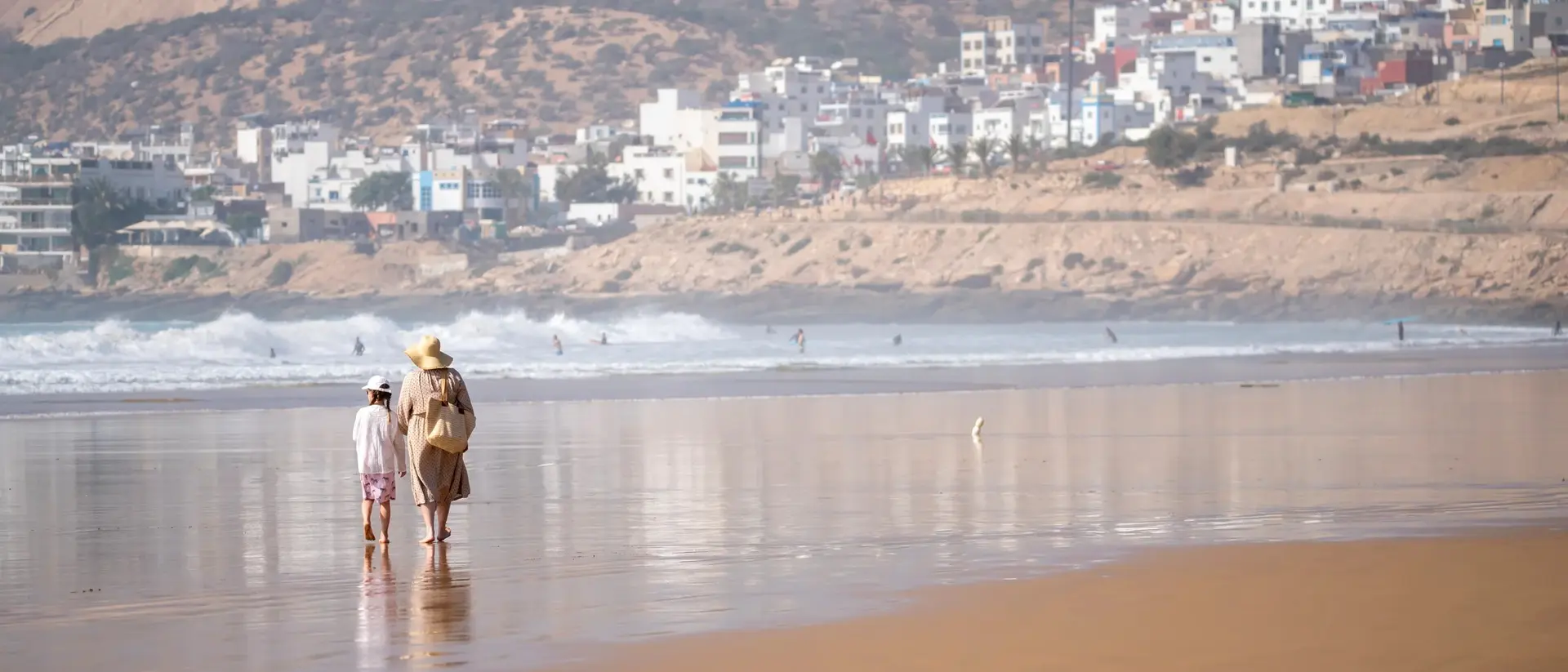 Two people walk along a sandy beach, with waves and a distant coastal town in the background.