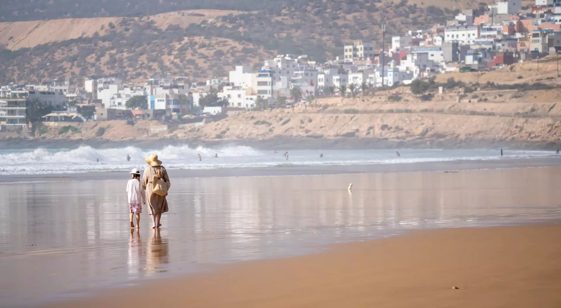 Two people walk along a sandy beach, with waves and a distant coastal town in the background.