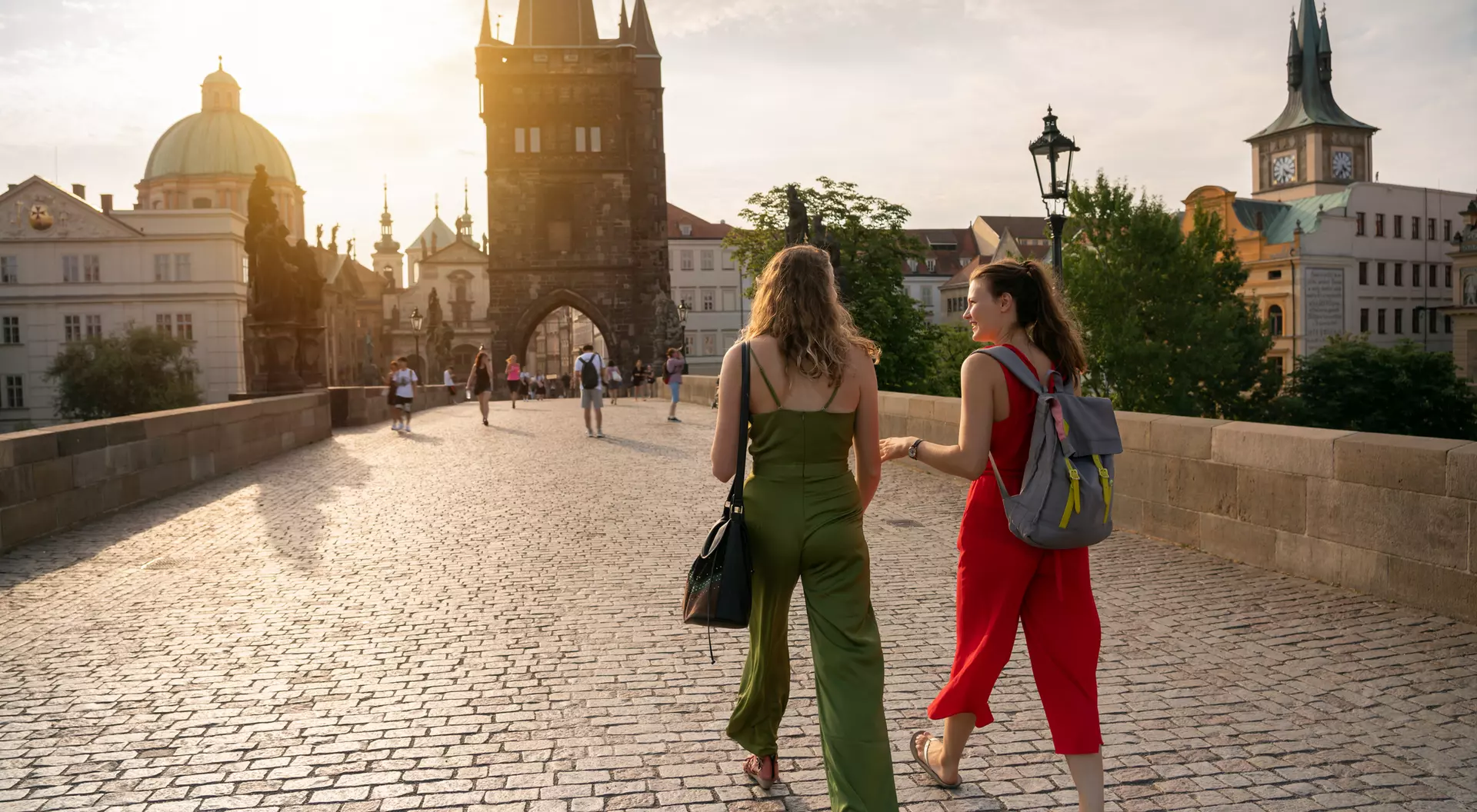 Two women stroll on a cobblestone bridge with historical buildings in the background during sunset.