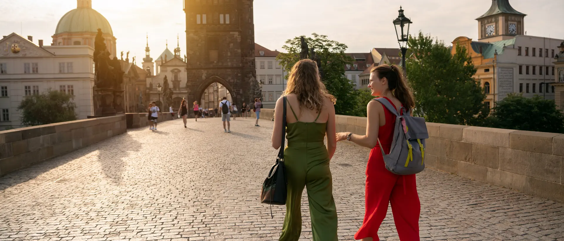 Two women stroll on a cobblestone bridge with historical buildings in the background during sunset.